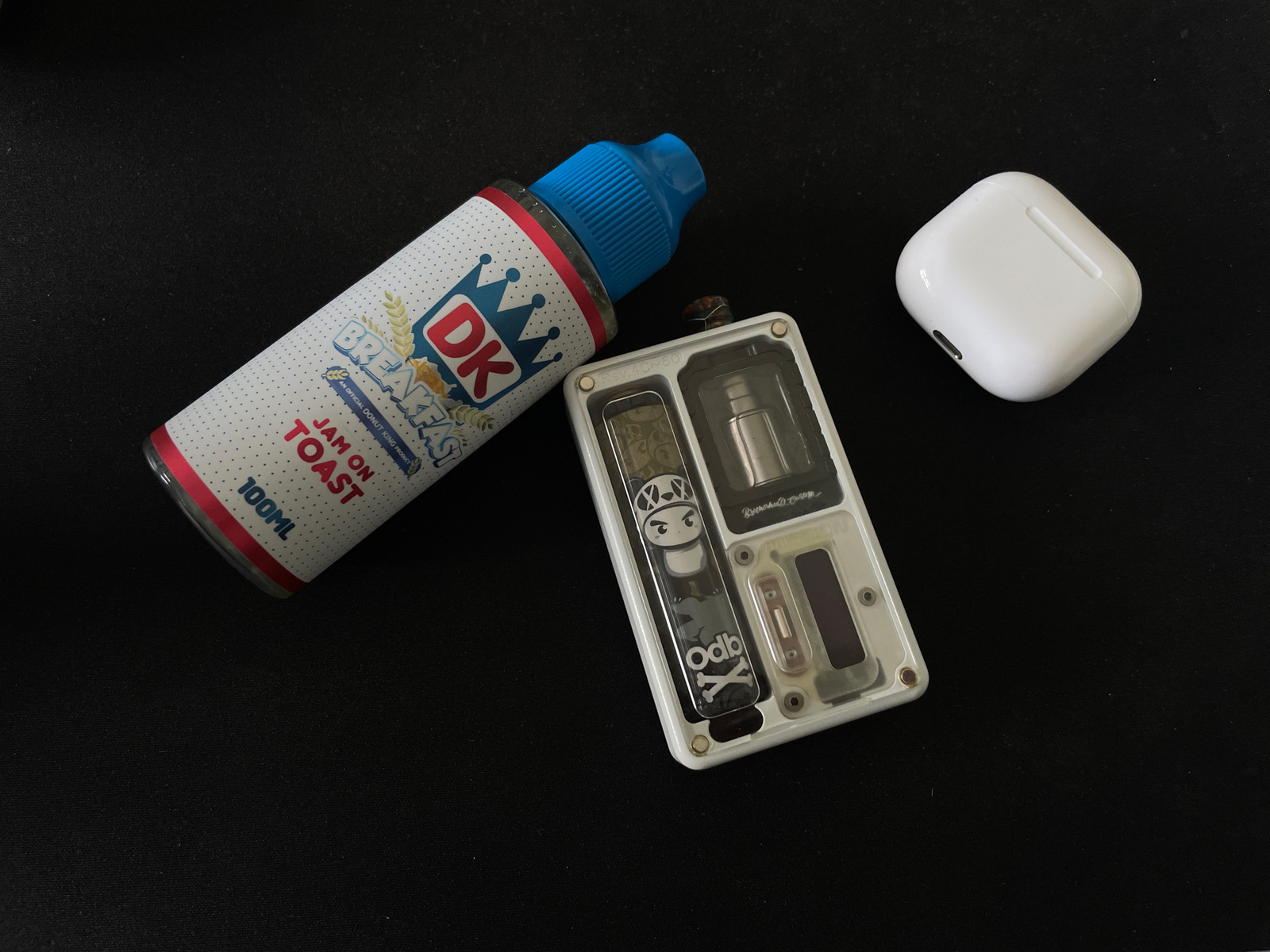 An overhead shot shows a black surface with three electronic items arranged in a triangular formation. On the left, a white bottle with a blue cap and a label featuring a crown and "DK BREAKFAST" is angled diagonally. The label also reads "JAM ON TOAST" and "100ML". In the center, a white rectangular vaping device with a clear front panel is positioned vertically. Visible through the panel are metallic components and decorative elements, including a cartoon character and the text "Odb X". To the right, a white, square wireless earbud charging case is placed. The environment appears to be indoors, with a dark, textured surface that could be a desk or cloth mat. There are no people or visible signs of activity. The image does not contain any information about the city of Voskresensk, Russia.