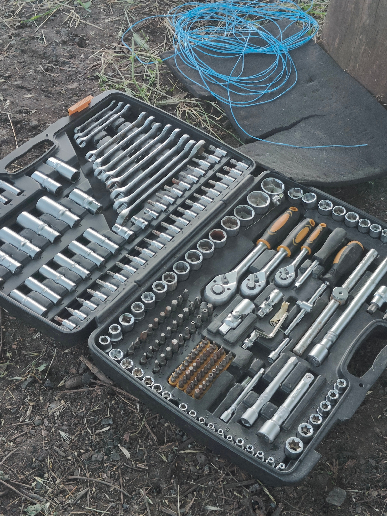A black plastic toolbox containing various mechanic tools, including wrenches, sockets, and ratchets, is open and lying on the ground. The ground appears to be dirt and debris, with dry grass and twigs. In the upper right corner of the image, a coil of thin blue wire is resting on a dark, flat object, possibly a piece of foam or rubber. The toolbox is organized with tools neatly placed in designated slots. The scene is outdoors, and given the ground cover and the presence of tools, it suggests a work area or storage location in Kryvyi Rih, Ukraine.