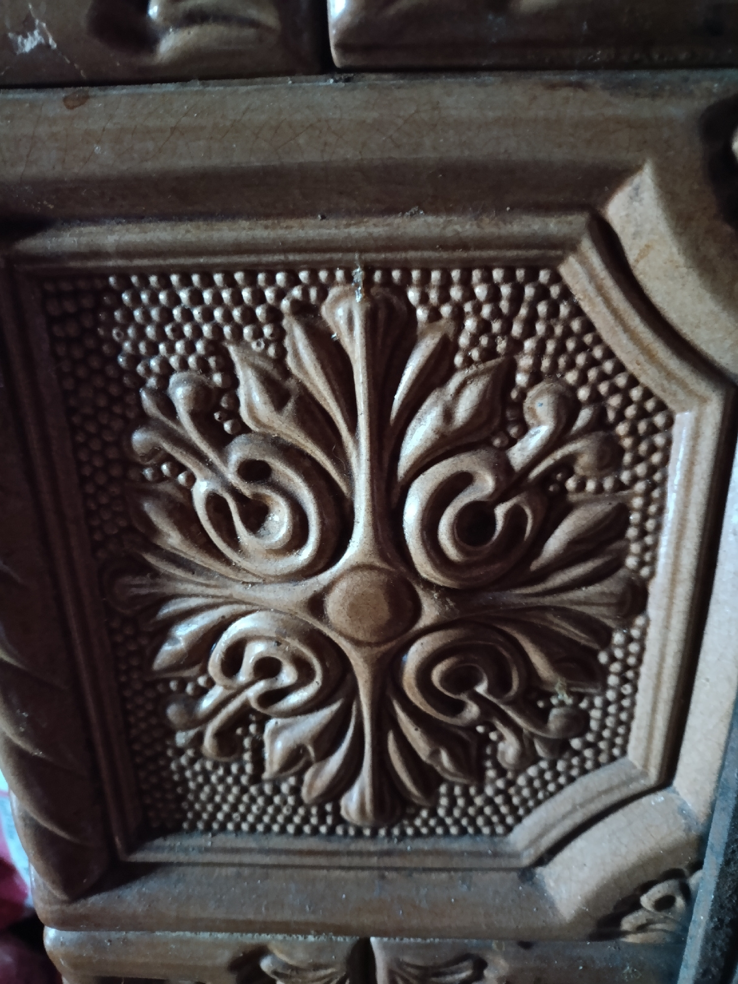 This is a close-up, high-angle, outdoor, natural light photograph of a carved, decorative ceramic tile. The tile has a raised, ornate design featuring stylized floral or leaf motifs and a textured background of small dots. The color of the tile is a muted, earthy brown with some variations in tone and subtle reflections on the glazed surface. The tile appears to be part of a larger structure or object, as indicated by the edges of surrounding elements visible in the frame. There are no people, animals, or other living subjects present. The background is blurred and suggests an indoor or outdoor setting with ambiguous elements, possibly part of a wall, furniture, or architectural feature. The overall impression is one of detailed craftsmanship and aged material.
