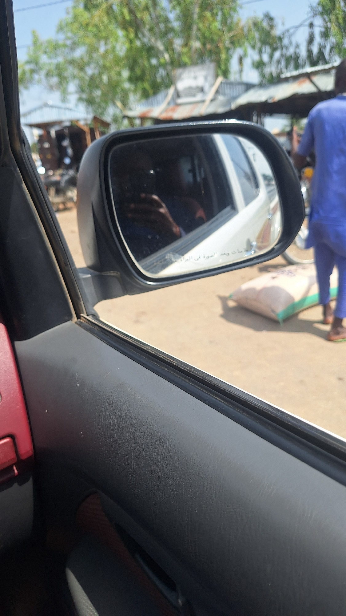 The image shows a view from inside a car, looking out the side window and into the side mirror. The side mirror reflects a person in a blue outfit, possibly a driver or passenger, and a white vehicle. Outside the car, there are several open-air structures with corrugated metal roofs, suggesting a market or roadside stalls. A large sack, light in color and with a green trim, is visible on the ground near a person wearing a blue shirt and pants. The ground appears to be dirt or gravel. Trees with green foliage are present in the background under a clear sky. The scene is likely a street or pathway in a rural or developing area.