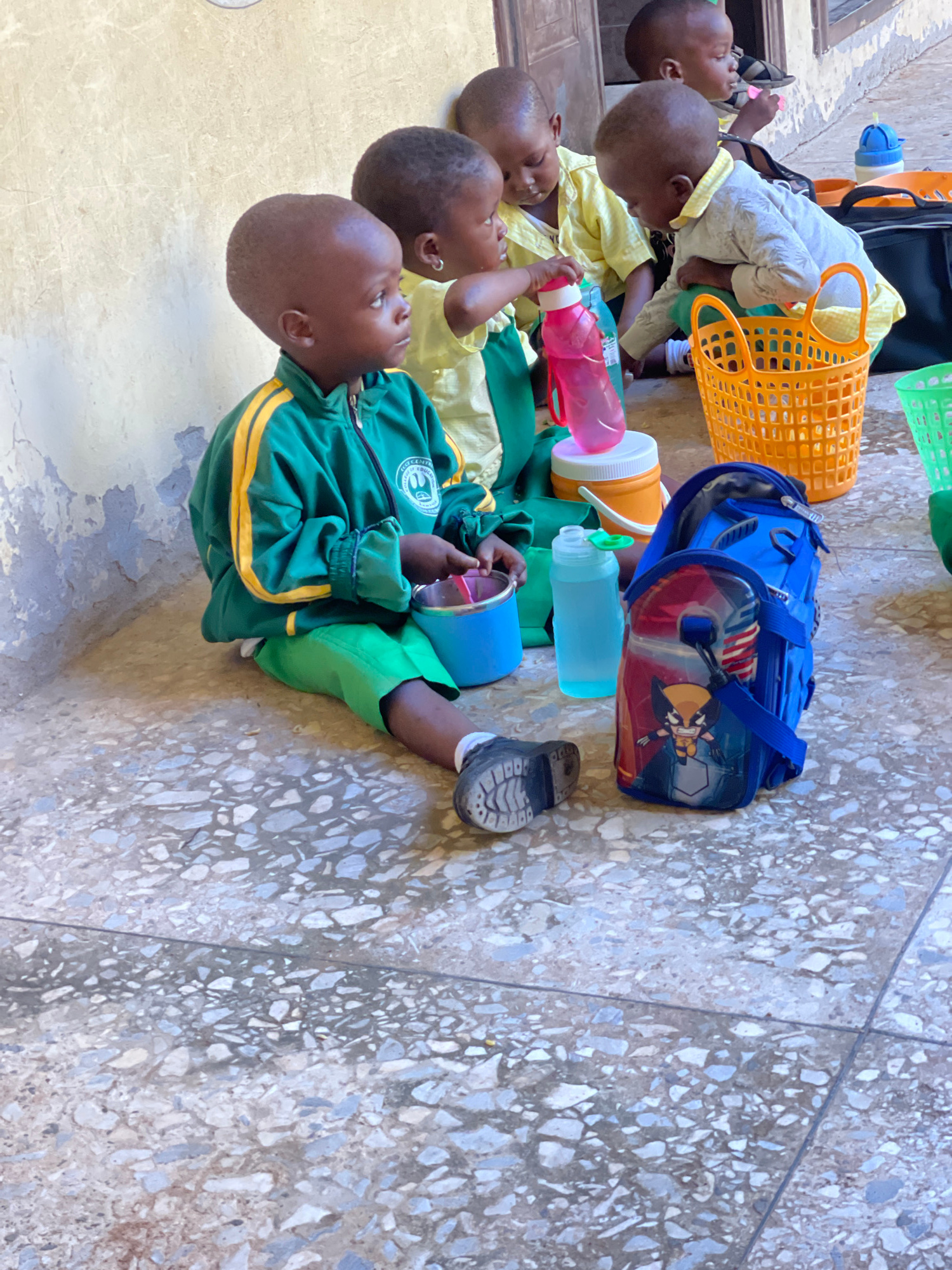 In Akwanga, Nigeria, a group of young children, likely preschoolers, are gathered outdoors on a terrazzo floor. The child in the foreground wears a green and yellow school uniform and is seated on the ground, looking to the right. In front of him is a blue lunchbox with a Wolverine graphic and a blue water bottle. To his right, an orange basket is visible, containing what appears to be other children's belongings. Behind him, other children in similar uniforms are seated, engaged in various activities, some holding water bottles and others appearing to eat. The environment suggests a school or community center setting, with a weathered wall in the background.