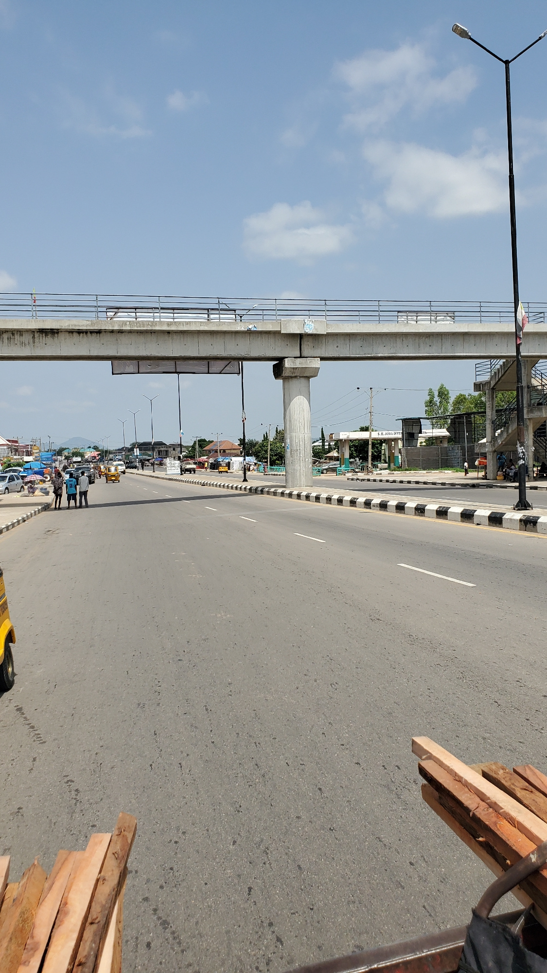 An elevated concrete road structure stretches across the frame, supported by large concrete pillars. Beneath it, a multi-lane asphalt road is visible, marked with white lines and a black and white striped border. Several vehicles, including cars, a tricycle-like vehicle, and a yellow auto-rickshaw, are present on the road. Pedestrians are seen on the sidewalk and near a building on the right. Wooden planks are piled on the lower right corner of the image, suggesting a construction or transport activity. The sky is blue with scattered white clouds. The scene appears to be a busy urban area in Jalingo, Nigeria, with a mix of modern infrastructure and everyday life.