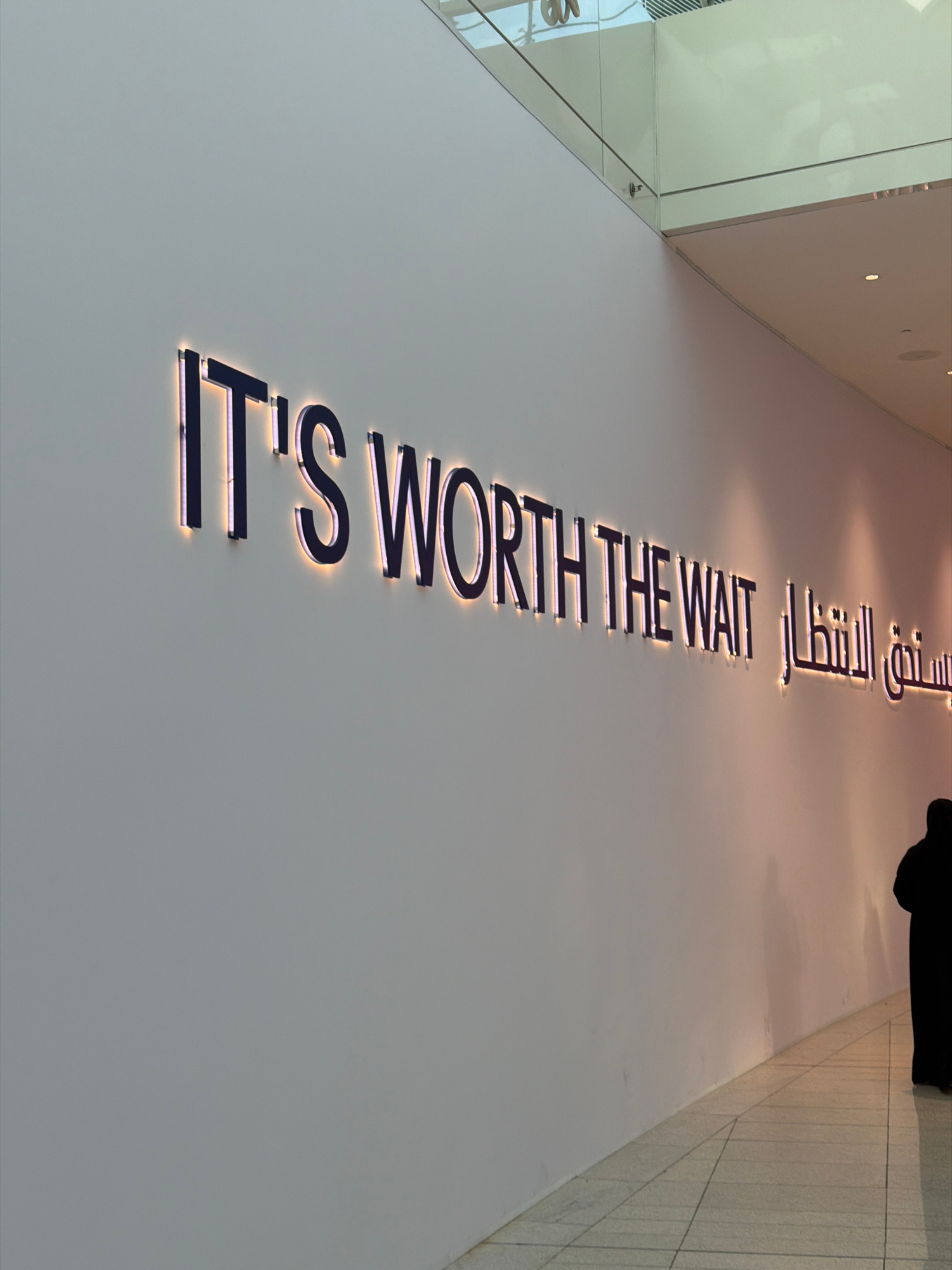 A long, off-white wall is illuminated by a bright, linear light source that casts a warm glow on the surface. Mounted on the wall are large, dark, three-dimensional letters that spell out "IT'S WORTH THE WAIT" in English. To the right of the English text, Arabic script with a similar illuminated effect is also present. In the lower right quadrant of the image, a person wearing a black robe is visible from the back, walking away from the camera down a corridor. The corridor floor is tiled with a grid pattern. Above the wall, architectural elements of a building are visible, including glass panels and a light-colored ceiling.