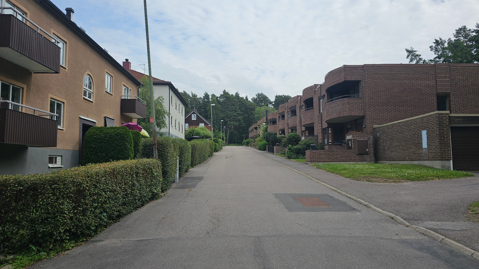The image depicts a quiet, suburban street lined with residential buildings and lush greenery. On the left, a light brown apartment building features multiple balconies with dark brown railings and a pitched roof with a red chimney. Adjacent to it, a white building with a darker roof is partially visible. The front of both buildings is bordered by a neatly trimmed green hedge.
On the right side of the street, a series of connected brick apartment buildings dominate the view. These buildings have a distinctive curved architectural style with balconies and dark windows. A low brick wall separates the buildings from the sidewalk, which is lined with grass and small white flowers.
The street itself is paved with asphalt and appears to be a dead end or leading into a more residential area, as it curves gently uphill. There are streetlights visible along the road. In the distance, a dense line of tall pine trees forms a natural backdrop to the neighborhood.
The sky is overcast with scattered clouds, suggesting a mild, possibly humid, day. The lighting is diffused, indicating it's likely daytime, perhaps late morning or afternoon. No people are visible in the image, and the scene conveys a sense of tranquility and suburban life. A small red sign with white text, partially obscured by a pole and greenery, is visible on the left side of the road. There's also a red square marking on the road on the right side. The overall impression is one of a well-maintained residential area in a temperate climate.