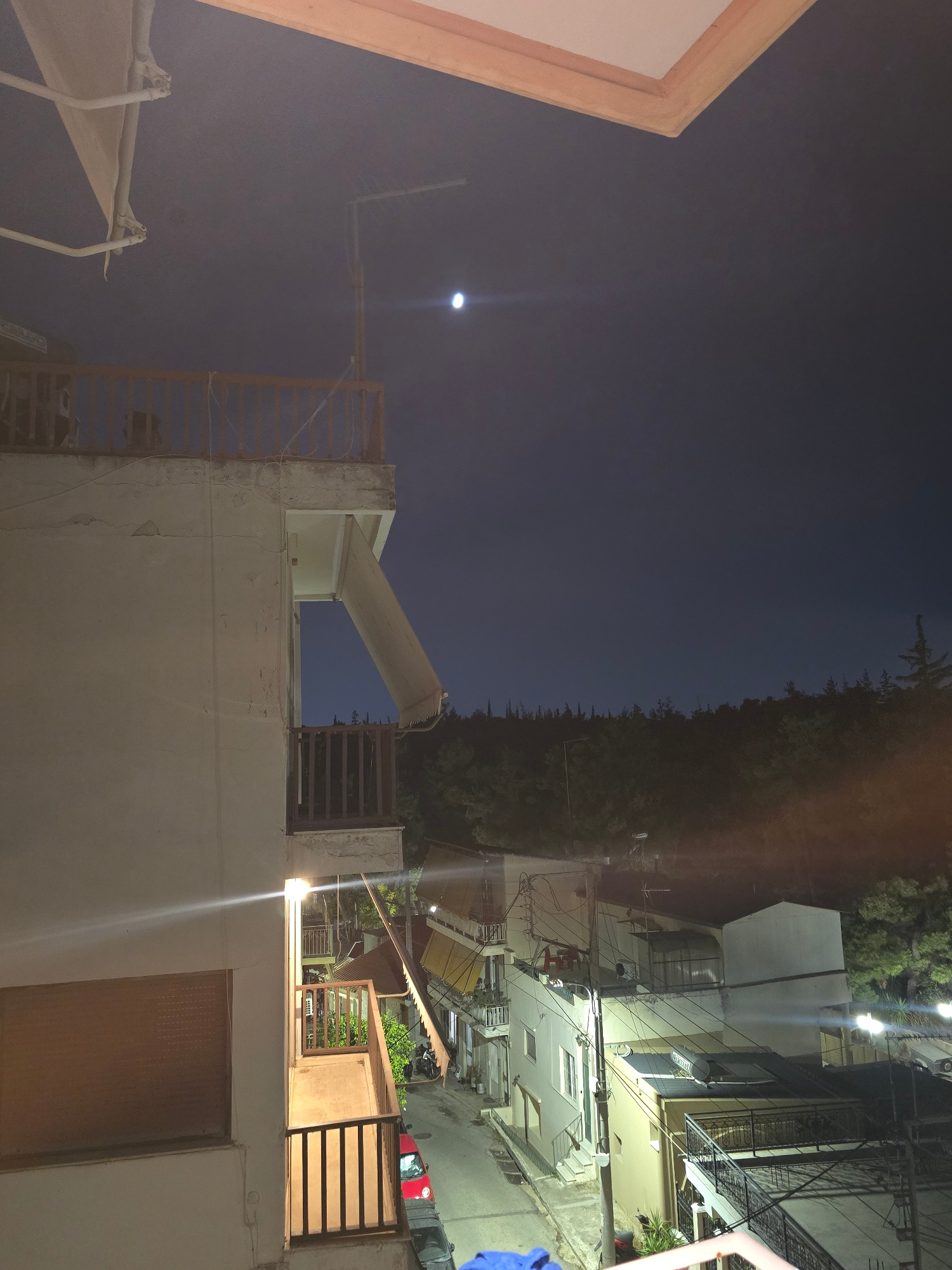 The image captures a nighttime view from an elevated vantage point, likely a balcony, in Nea Ionia, Greece. The scene is dominated by the dark blue of the night sky, with a bright, full moon visible near the top center. Below the sky, a dense treeline with visible conifer trees forms the backdrop to a residential area.
In the foreground and midground, multi-story buildings are visible, characterized by their apartment-like structures and balconies. One building on the left is prominently featured, showing a white-rendered facade with balconies and railings. A retractable awning is partially extended from one of the upper balconies. Several other buildings with similar architectural styles, including balconies, awnings, and staircases, are visible in the midground, suggesting a dense urban or suburban setting.
The street level below reveals a red car parked on the asphalt road, with a motorcycle also present. Streetlights cast pools of light, illuminating parts of the street and the lower levels of the buildings. The overall impression is one of a quiet residential neighborhood at night, with the natural element of the treeline providing a contrast to the urban environment.