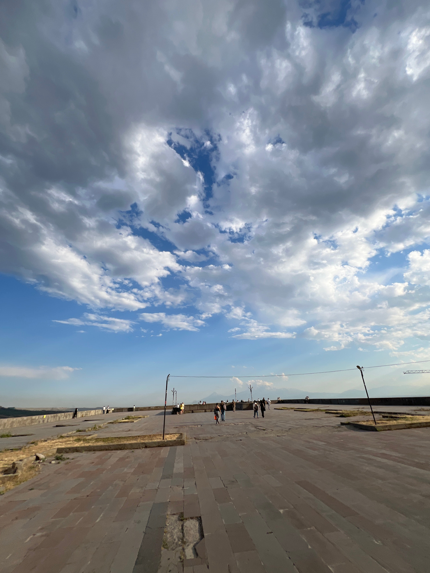 A wide-angle, low-angle shot captures a paved plaza under a dramatic, cloudy sky. The plaza, made of large, rectangular stone tiles, stretches into the distance. Patches of dry grass and small, square garden beds are interspersed on the left side of the plaza. Several people are visible walking and standing along the far edge of the plaza, which is bordered by a low stone wall. Two tall, thin poles with wires strung between them are prominent features, one in the middle ground and another further to the right. In the background, a hazy landscape with distant mountains and a clear blue sky peeking through the clouds can be seen. The scene appears to be a public space in the city of Yerevan, Armenia, possibly the Cascade complex or a similar viewpoint.