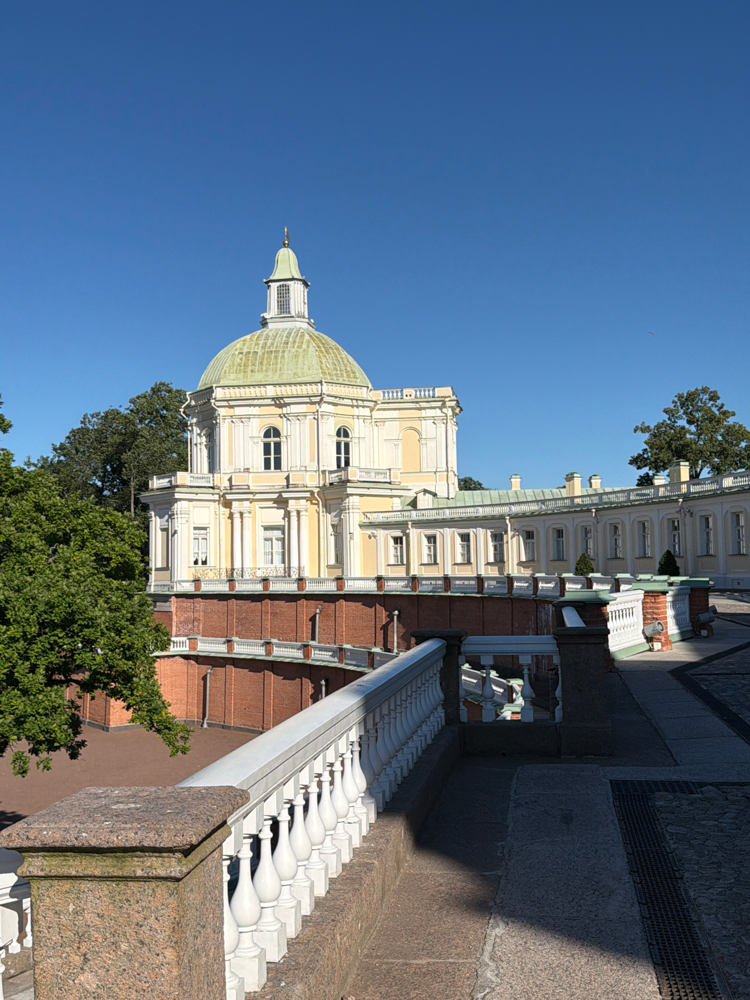 The image captures a grand architectural structure, likely a palace or museum, set against a clear blue sky. The building features a prominent green dome topped with a small spire. Its facade is a light yellow with white decorative elements, columns, and arched windows. A red brick base supports the main structure, which is further embellished with white balustrades and balconies. Lush green trees frame the left side of the building. In the foreground, a white balustrade runs along a granite-clad pathway, leading the eye towards the grand building. The pathway itself is paved with stones and features a metal grate. The scene is bathed in sunlight, casting distinct shadows on the ground. There are no people or animals visible in the image.