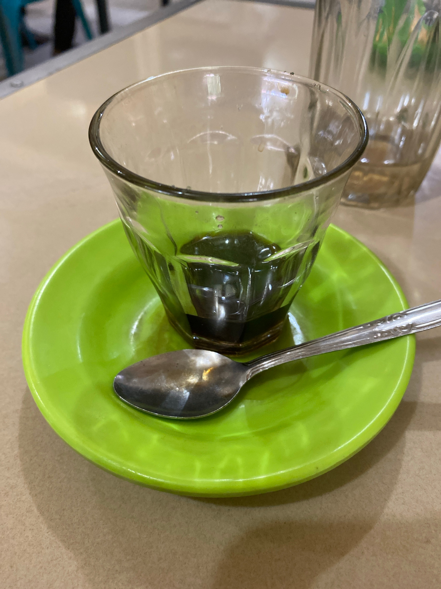 A close-up, eye-level shot shows a small, clear glass with a dark liquid, likely coffee, sitting on a bright green saucer. A silver spoon rests on the saucer in front of the glass. The background is blurred, but hints at a table and other glasses, suggesting an indoor setting like a cafe or restaurant in Lhokseumawe, Indonesia.