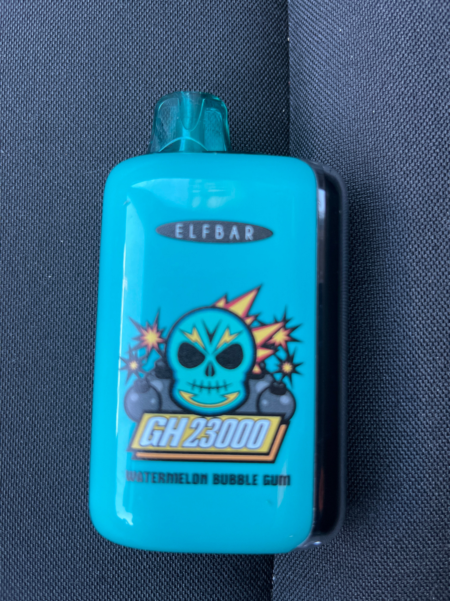 The image shows a bright teal disposable electronic cigarette device resting on a dark, textured surface, likely fabric. The device is rectangular with rounded edges and features a black top and a translucent teal mouthpiece extending from the left side.
Prominently displayed on the device is a graphic of a stylized skull with a turquoise face, black eyes, and teeth. Explosions with yellow and orange starbursts emanate from behind the skull. To the right of the skull, in a yellow rectangular box with a black outline, is the text "GH 23000". To the left of the skull, vertically oriented within a black oval shape, is the text "ELFBAR". Further to the right, also vertically oriented, is the text "WATERMELON BUBBLE GUM".
The background is a uniform dark color with a distinct woven texture, suggesting it is a piece of furniture or upholstery. The lighting appears to be natural daylight, casting a soft, even illumination on the object. No people are visible in the image. The scene is static, with the electronic cigarette as the sole subject. The location is identified as Kharkiv, Ukraine, though no specific visual cues within the image confirm this. The overall impression is a close-up product shot.