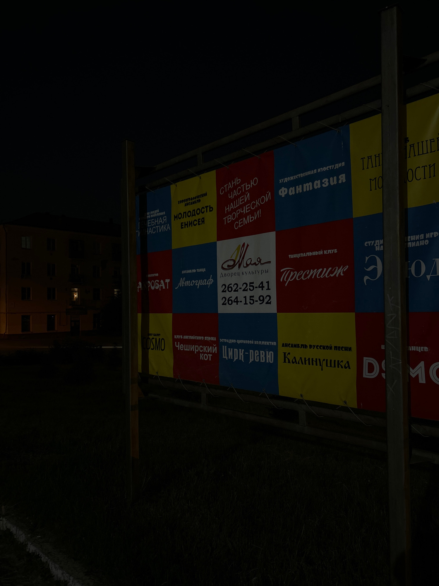 The image shows a brightly colored banner with various advertisements for different local establishments, displayed outdoors at night. The banner is mounted on a metal frame and appears to be positioned in front of a dark, grassy area with a few lights visible in the distance, suggesting an outdoor venue or open space. The lighting is dim, typical of nighttime, and the banner itself is illuminated, making the text and graphics stand out.
The banner is divided into several sections, each promoting a different service or group. Text in Russian is prominently displayed, advertising things like a "Choreographic Ensemble," "Art Studio," "Dance Club," "Beauty Studio," "English Language Club," "Russian Song Ensemble," and a "Circus-Review." Specific names like "Fantasia," "Prestige," "Kalininushka," and "Cheshirsky Kot" are visible. Phone numbers are also present, such as "262-25-41" and "264-15-92." The overall impression is one of community advertising, likely for local entertainment and cultural activities. The bright colors of the banner contrast with the dark surroundings, drawing attention to the advertisements. The background is a dark, out-of-focus scene of what appears to be a building with lit windows and a slightly visible, overgrown grassy area with some faint markings.