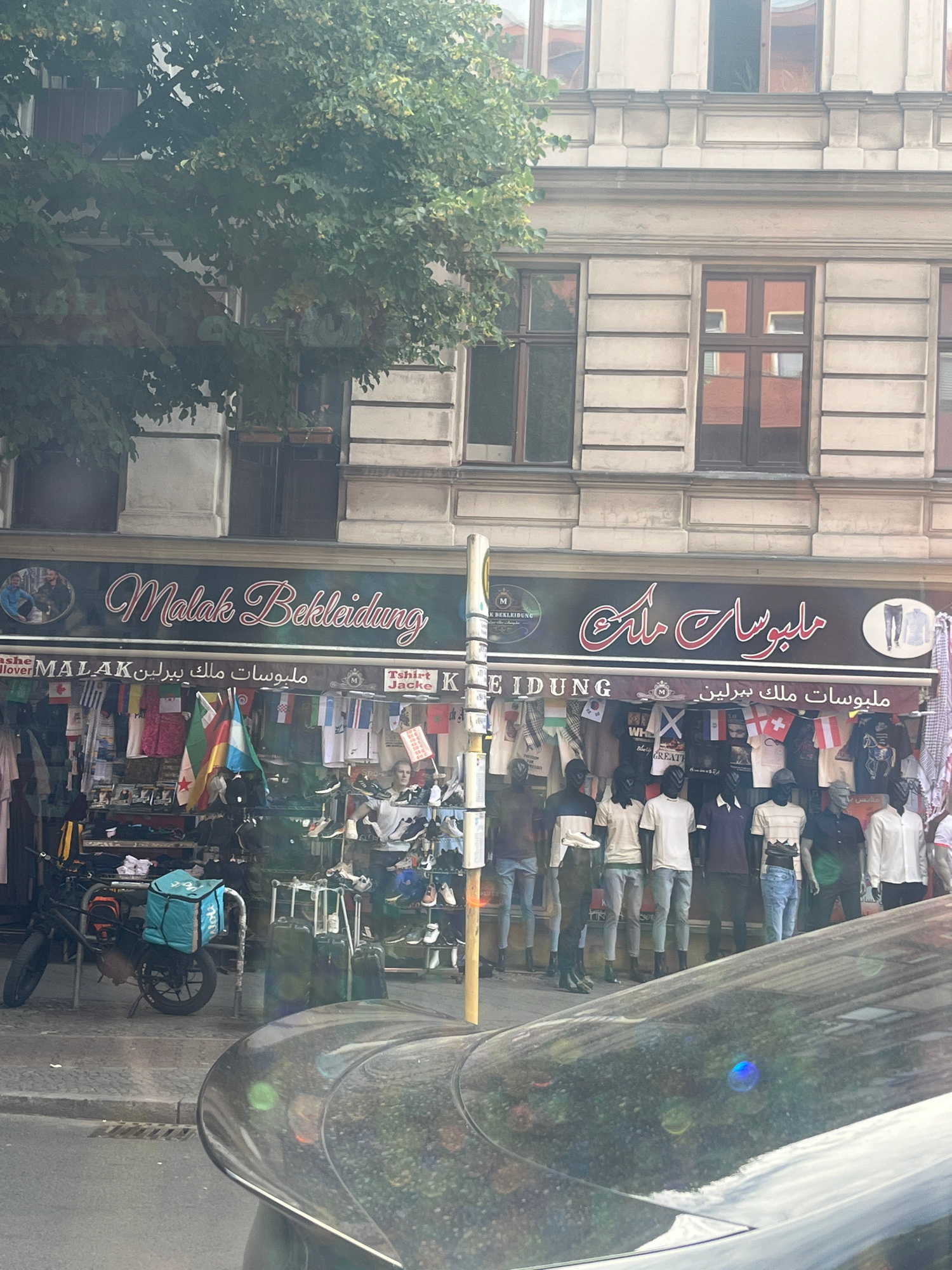 This is a medium-shot, eye-level, outdoor photograph taken from a car, showing the facade of a clothing store called "Malak Bekleidung". The store is located on a street in Berlin, Germany, as indicated by the street names on the awnings and the German word "Bekleidung" (clothing). The store features a prominent dark awning with the store name in both Latin and Arabic script. Below the awning, the storefront is filled with clothing and mannequins displayed for sale. Flags of various countries are hung across the front of the store. To the left of the store, a delivery bicycle with a blue delivery bag is parked. The building itself is a multi-story structure with architectural details typical of Berlin. The reflection on the car in the foreground obscures some of the scene.