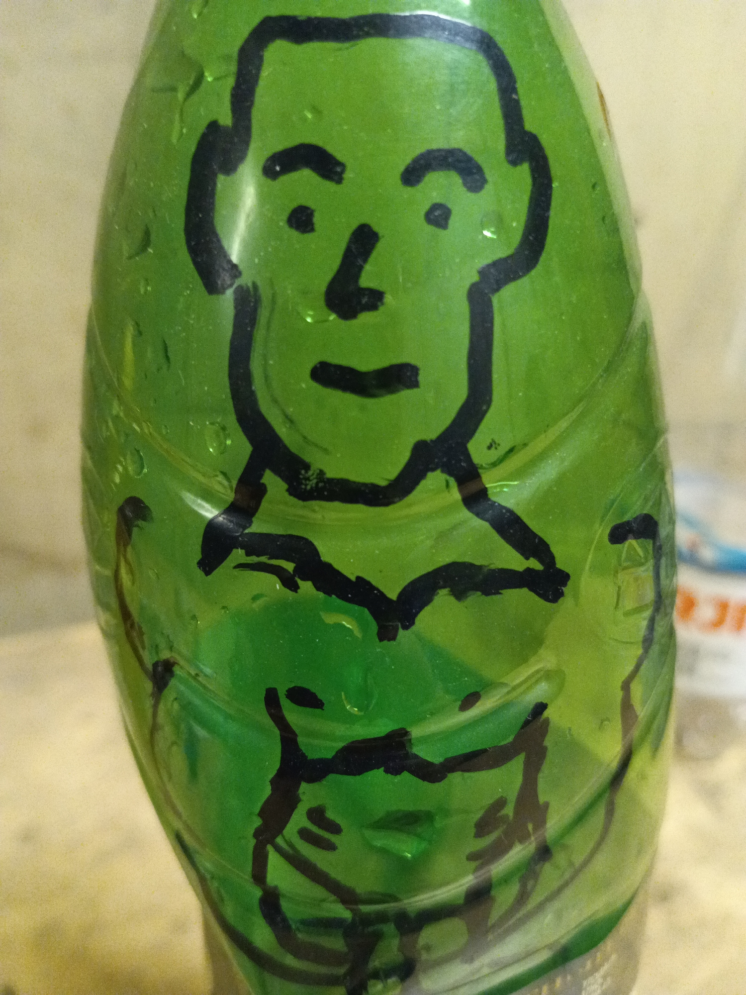 A close-up, vertical shot focuses on a green plastic bottle that has been decorated with a black marker. The marker art depicts a stylized human face at the top of the bottle, with simple black lines for eyes, nose, mouth, and a head outline. Below the face, more black marker lines suggest clothing or a torso. The bottle is slightly curved and appears to have water droplets on its surface, indicating condensation. The background is blurred and appears to be a light-colored, textured surface, possibly a table or countertop. In the far background, some out-of-focus elements suggest other objects or a wall, with hints of color that are difficult to discern clearly. The lighting suggests an indoor setting, possibly with artificial light. No specific time of day or weather conditions can be inferred. The location context is Saint Petersburg, Russia, but there are no explicit visual cues within the image to confirm this. No legible text is visible on the bottle or in the surrounding environment. The overall impression is of a simple, whimsical decoration applied to a common object.