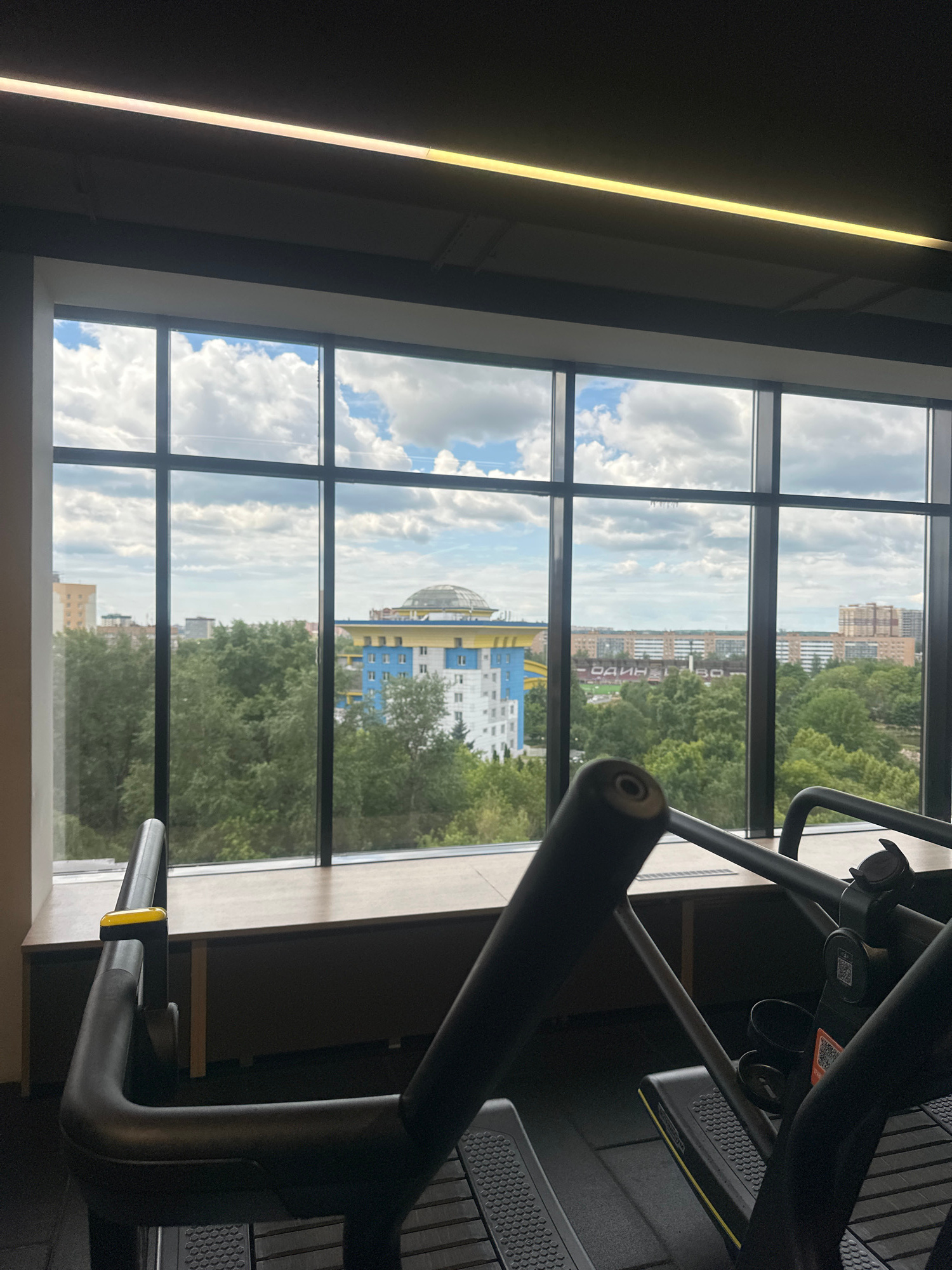 An interior shot shows a gym with a large window overlooking a city skyline. In the foreground, the handles and part of the frame of an elliptical machine are visible. The window is divided into multiple panes by black mullions, and through it, one can see a variety of buildings and trees. The sky is partly cloudy with white clouds against a blue backdrop. Some of the buildings are residential, with several stories and apartment-style windows. One building has a distinctive, domed roof structure. The overall impression is a modern fitness facility with an expansive view of an urban environment.