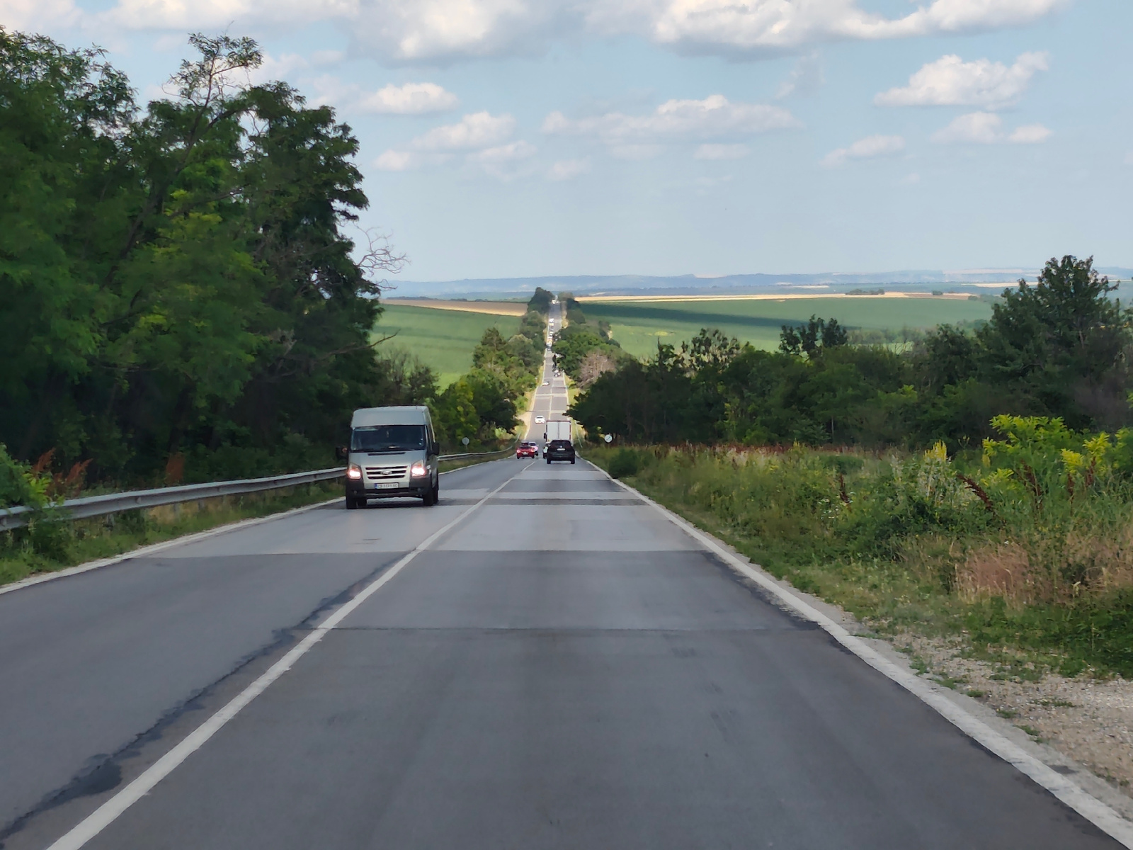 The media file depicts a highway scene in Zgalevo, Bulgaria, under a partly cloudy sky. The time appears to be late afternoon, with soft sunlight casting shadows. The road, a two-lane asphalt highway, stretches into the distance, lined by lush green trees on both sides. In the foreground, a silver van is driving on the right side of the road. Further down the highway, several other vehicles, including a white truck, a red car, and a dark-colored car, are visible. The landscape beyond the trees opens up to rolling fields, some appearing green and cultivated, while others are a lighter, possibly harvested, golden hue. The distant horizon shows a faint range of hills or mountains. The overall atmosphere is peaceful and rural, suggesting a typical day of travel through the Bulgarian countryside. The white line markings on the road are clear, indicating good visibility. No people are distinctly visible, but the presence of vehicles implies ongoing travel. The weather appears pleasant, suitable for driving and outdoor scenery. There is visible text on the license plate of the silver van, though it is not clearly legible.