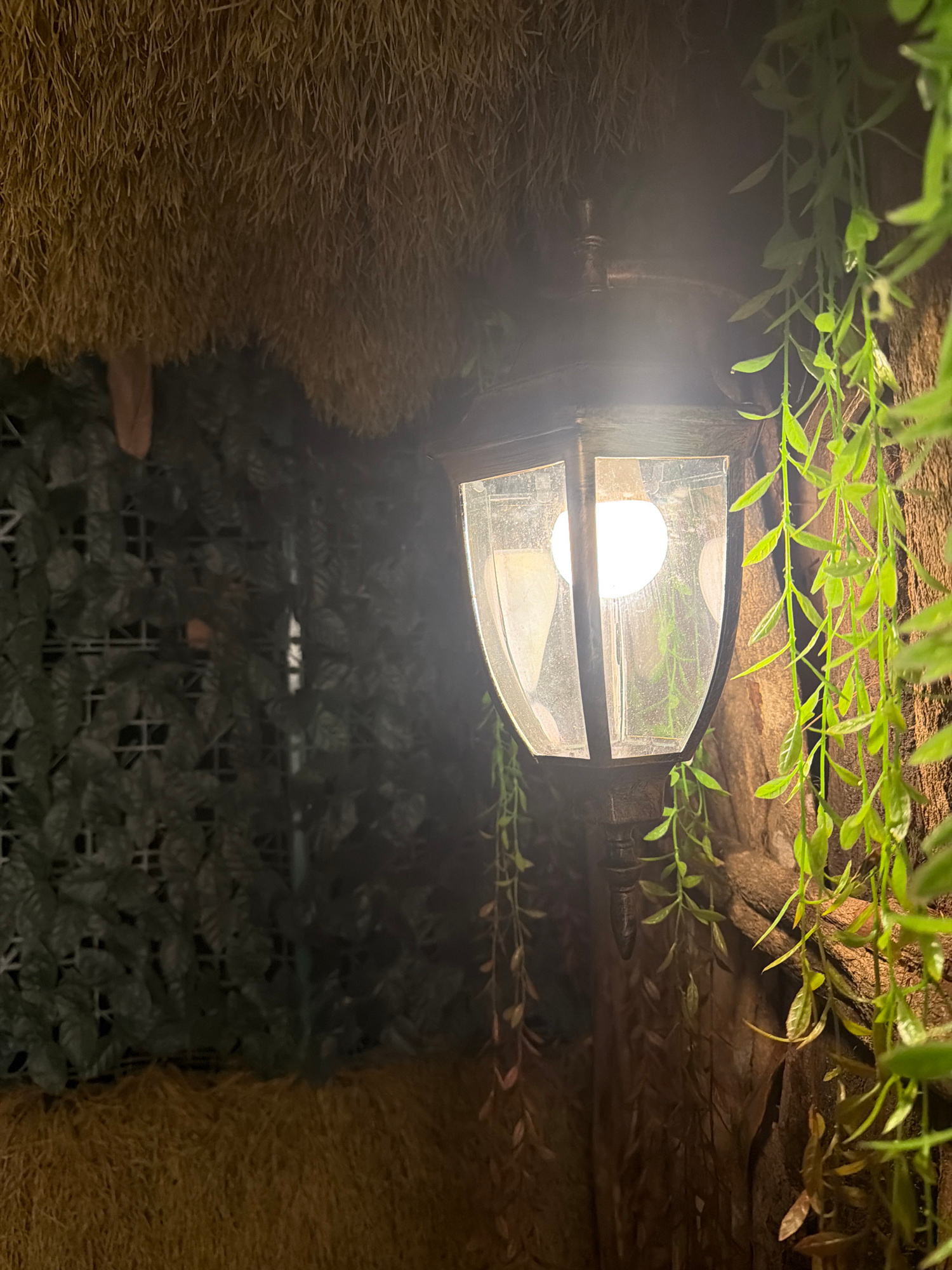 A close-up, vertical shot reveals a dimly lit, rustic setting, possibly a terrarium or a decorative enclosure. The primary object is an old-fashioned lantern with a transparent pane, emitting a soft, warm glow from an unseen light bulb within. The lantern is partially obscured by a tangle of artificial green foliage, which hangs from above and drapes around it. The background is a mix of textures: to the left, a coarse, straw-like material creates a textured wall. To the right and below, a dense covering of dark green artificial leaves is laid out over a grid-like structure, suggesting a wall or floor covering. The overall impression is one of an enclosed, artificial natural environment, likely designed for aesthetic appeal. No people are visible, and the lighting suggests it could be evening or a low-light interior. No text is visible.