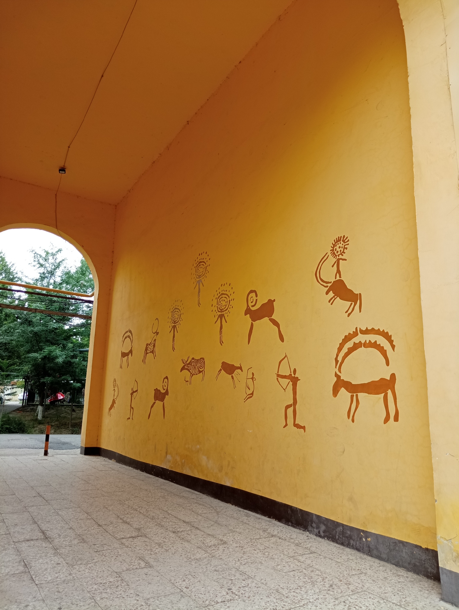 The image depicts a section of a building's ceiling decorated with stylized cave paintings. The paintings, rendered in a reddish-brown color, feature depictions of humans and animals, reminiscent of ancient rock art. Among the figures are what appear to be human figures in various poses, some with sun-like halos, and several animals including what look like horses or deer, and possibly a goat. One human figure is shown with a bow and arrow. The ceiling itself is painted a warm, yellowish-orange color and is illuminated by natural light, suggesting it is daytime. The setting appears to be an outdoor or semi-outdoor space, possibly an overhang or a covered walkway, given the presence of an archway in the lower left of the image, revealing trees and foliage beyond. The floor to the right is tiled with light-colored stone. The overall impression is one of cultural or historical decoration applied to a modern structure. There is no visible text in the image.
