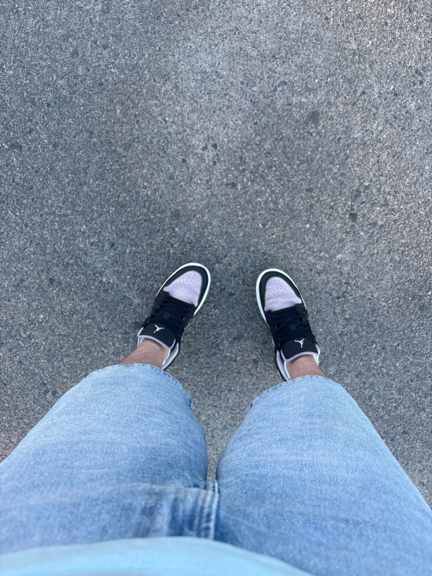 A first-person perspective, overhead shot of a person standing on a paved surface, likely asphalt, suggesting an outdoor urban environment in Dnipro, Ukraine. The individual is wearing light blue denim shorts and black and white sneakers with purple accents. The sneakers appear to be low-top athletic shoes. The ground is a consistent texture of dark gray asphalt with visible aggregate. The lighting suggests daytime.