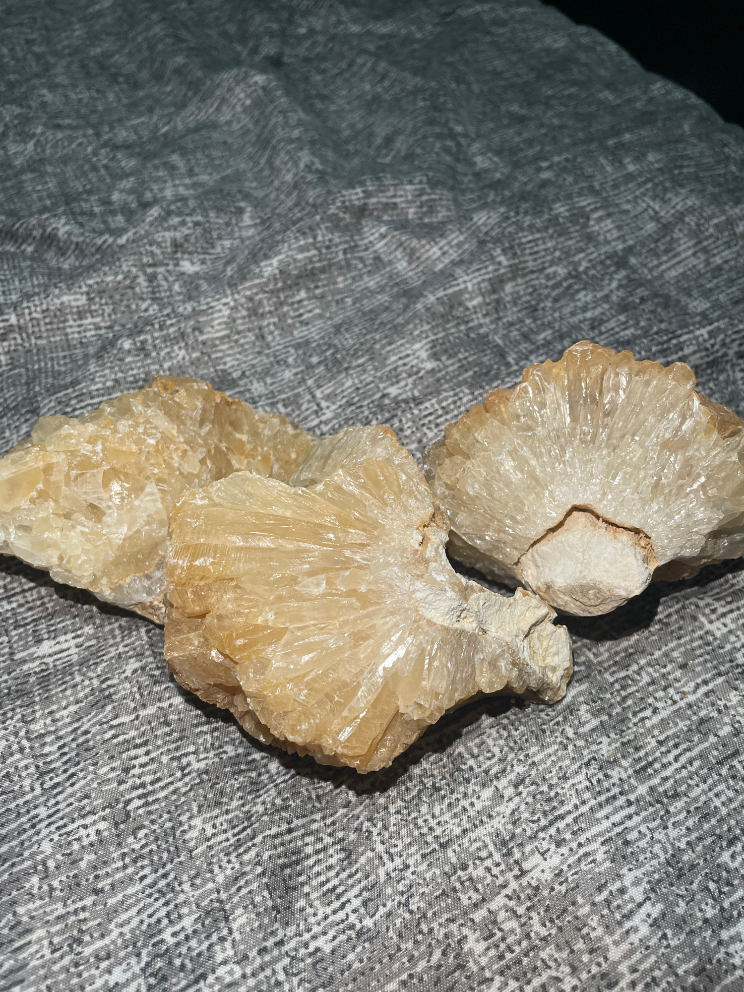 The image displays three light yellow, crystalline mineral specimens resting on a gray, textured fabric. The crystals exhibit a fan-like or radiating growth pattern, with many elongated, prismatic facets. The fabric has a subtle, woven pattern that appears as a series of short, parallel lines and dashes. The lighting casts shadows on the fabric, highlighting the texture and the forms of the mineral samples. There are no people or any indication of a specific location in Davao City, Philippines, visible in the image. The scene is an indoor still life arrangement of geological specimens.