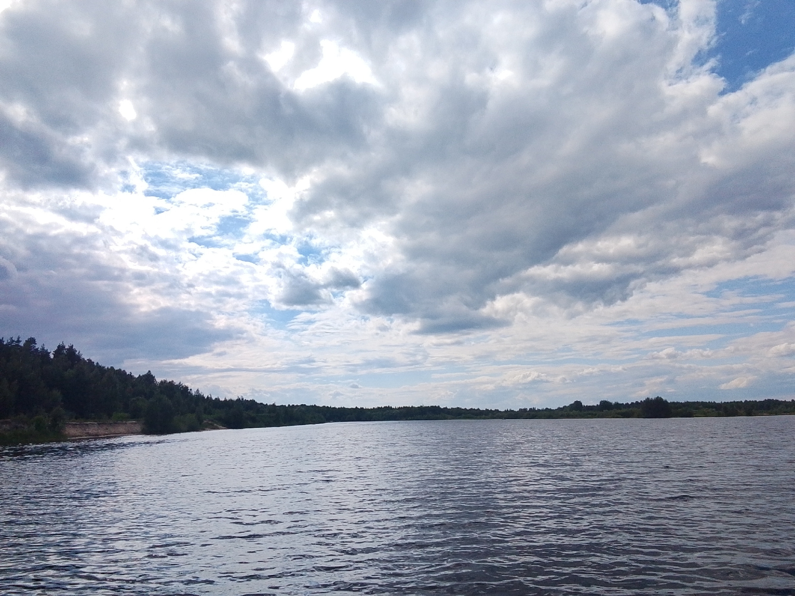 This media file depicts a wide, calm river under a dramatic sky filled with clouds. The river occupies the foreground and midground, its surface rippling slightly, reflecting the light from the overcast sky. On the left side, a densely forested shoreline with dark green trees stretches into the distance, meeting a sandy bank near the water's edge. The opposite shore is visible as a distant, continuous line of trees, suggesting a broad river or lake.
The sky is a prominent feature, dominated by a mix of dark, heavy clouds and patches of brighter blue sky where sunlight breaks through. The light filtering through the clouds creates a dynamic and somewhat moody atmosphere. The overall impression is of a natural landscape, likely captured on a day with varied weather. There are no people or man-made objects visible in the image. The scene appears to be set in the daytime, given the visible sky and lighting conditions. The location is not specified but appears to be a natural, possibly remote, area with a significant body of water and extensive woodland. No text is visible in the image.