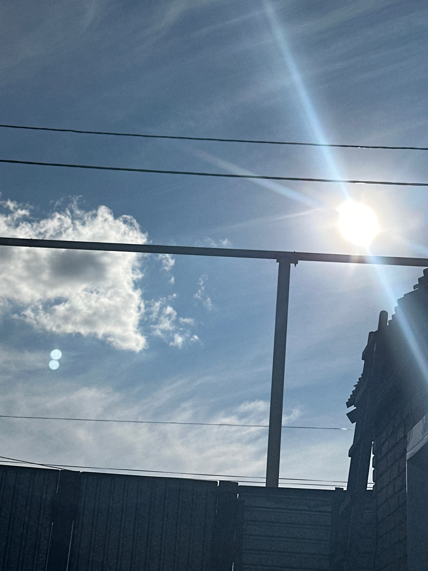 The image captures a bright, sunny day in the city of Tobyl, Kazakhstan. The sky is a vibrant blue, with thin, wispy clouds scattered across it. The sun is shining intensely, creating a strong lens flare that is visible in the upper right quadrant of the image. Below the sky, a dark, corrugated metal fence runs horizontally across the lower portion of the frame, suggesting an outdoor, possibly urban or industrial, setting. Two thin power lines stretch across the sky from left to right, adding a linear element to the composition. In the middle ground, a vertical metal pole supports a horizontal bar, which is likely part of a fence or structure. On the far right, the dark silhouette of a building's roofline is visible, its architectural details partially obscured by the bright sunlight. Two circular, lens-flare artifacts are present in the sky above the fence, appearing as bright, overlapping orbs. No people or animals are visible in the scene. The overall impression is one of a clear, sunny day with evidence of infrastructure and structures typical of a populated area.