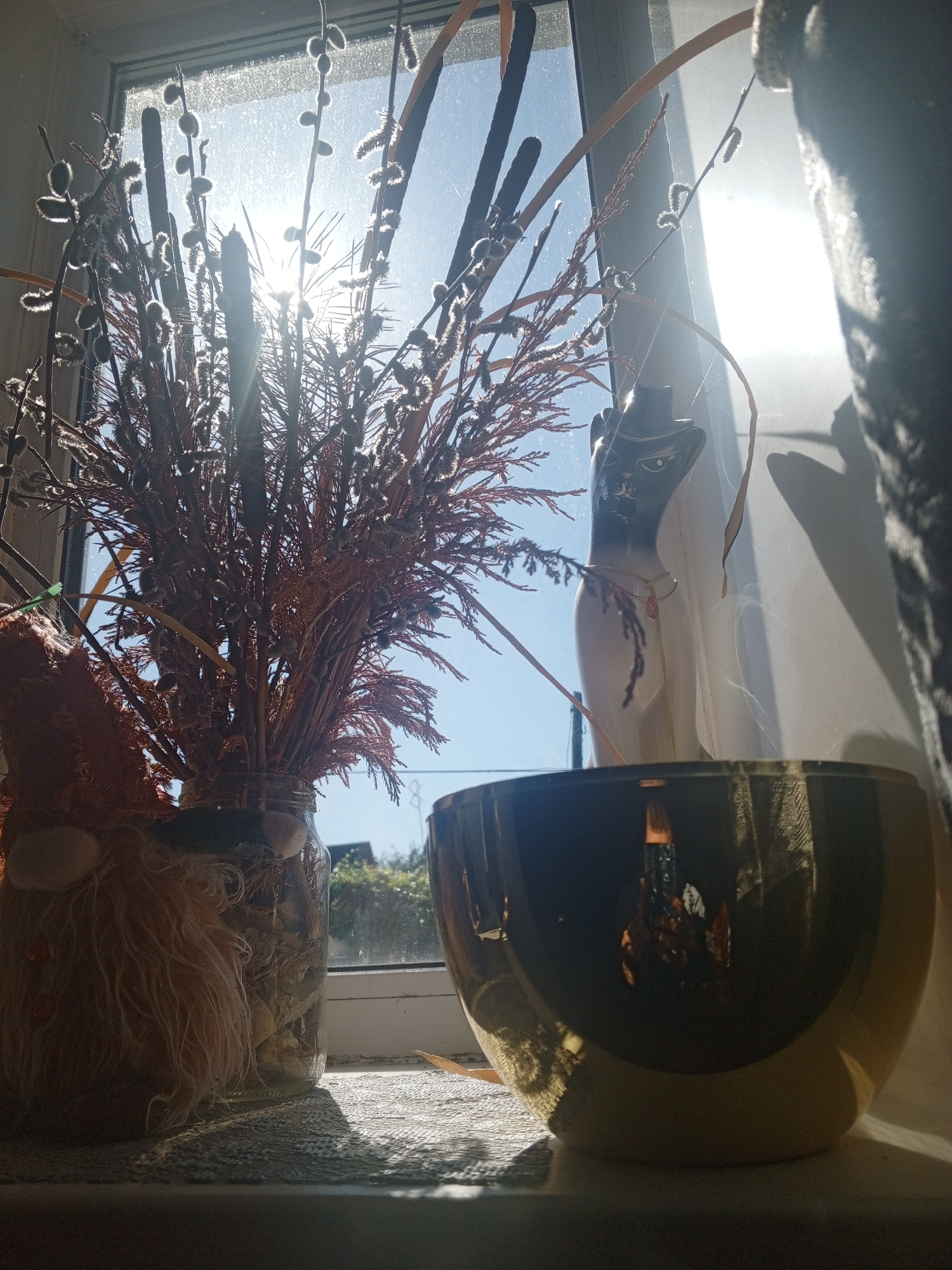 A still life composition is captured through a window with bright sunlight streaming in. In the foreground, a large, gold-toned bowl reflects a darker interior, casting shadows on a textured surface. To the left, a glass jar holds a bouquet of dried reeds and pussy willows, some with fluffy white catkins. This jar is partially obscured by a decorative object on its left, which appears to be a gnome or similar figure with a shaggy, light-brown beard. Behind the jar and bouquet, a white ceramic figurine shaped like a cat wearing a black hat with a gold trim stands next to the window. The figurine is adorned with a thin necklace featuring a pink pendant. The background consists of the window pane, through which a bright blue sky and some distant green foliage are visible. The sunlight creates lens flare and highlights, obscuring some details and casting strong shadows. The overall scene is set indoors, on a windowsill or ledge. The location is stated as Saltykovka, Russia, though no specific city landmarks are visible to confirm this.