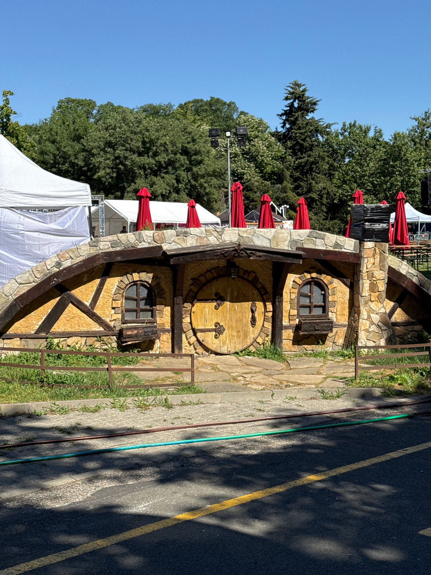 The image captures a whimsical, hobbit-hole-inspired structure situated outdoors, likely at an event or park. The structure features a rounded, wooden door with decorative metal hardware, flanked by two small, arched windows. The facade is a mix of rough-hewn stone and textured yellow plaster, with dark wooden beams forming decorative accents. A stone archway curves over the entrance, adorned with several closed, bright red umbrellas. Behind the structure, white tents and a stage setup are visible, suggesting an outdoor festival or gathering. Trees form a dense backdrop under a clear blue sky. A low, rusty metal fence runs in front of the structure, separating it from a paved walkway and a road where two hoses, one green and one brown, lie on the asphalt. The scene is bathed in bright sunlight, casting distinct shadows. This structure is located in the Sea Garden park in Burgas, Bulgaria, a popular public space often used for events and leisure activities.