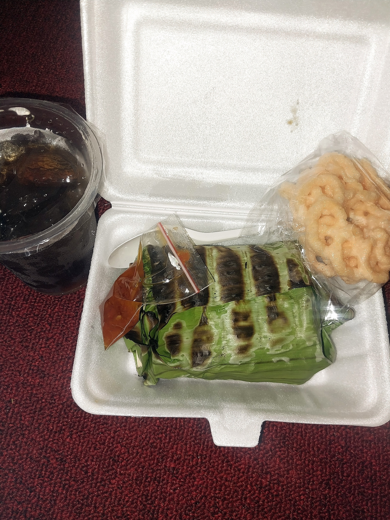 A Styrofoam takeout container is placed on a dark red, textured surface, possibly carpet. The container holds a meal wrapped in banana leaves. Some of the banana leaves show signs of being charred or grilled. A small plastic bag containing a reddish sauce is also visible, partially covering the food. To the right of the food, another plastic bag contains a light-colored, puffed snack, possibly crackers. To the left of the container, a clear plastic cup is filled with a dark liquid, likely a beverage. The scene is lit from above, casting shadows. The image does not provide specific visual cues to determine its exact location within Tibojong, Indonesia.