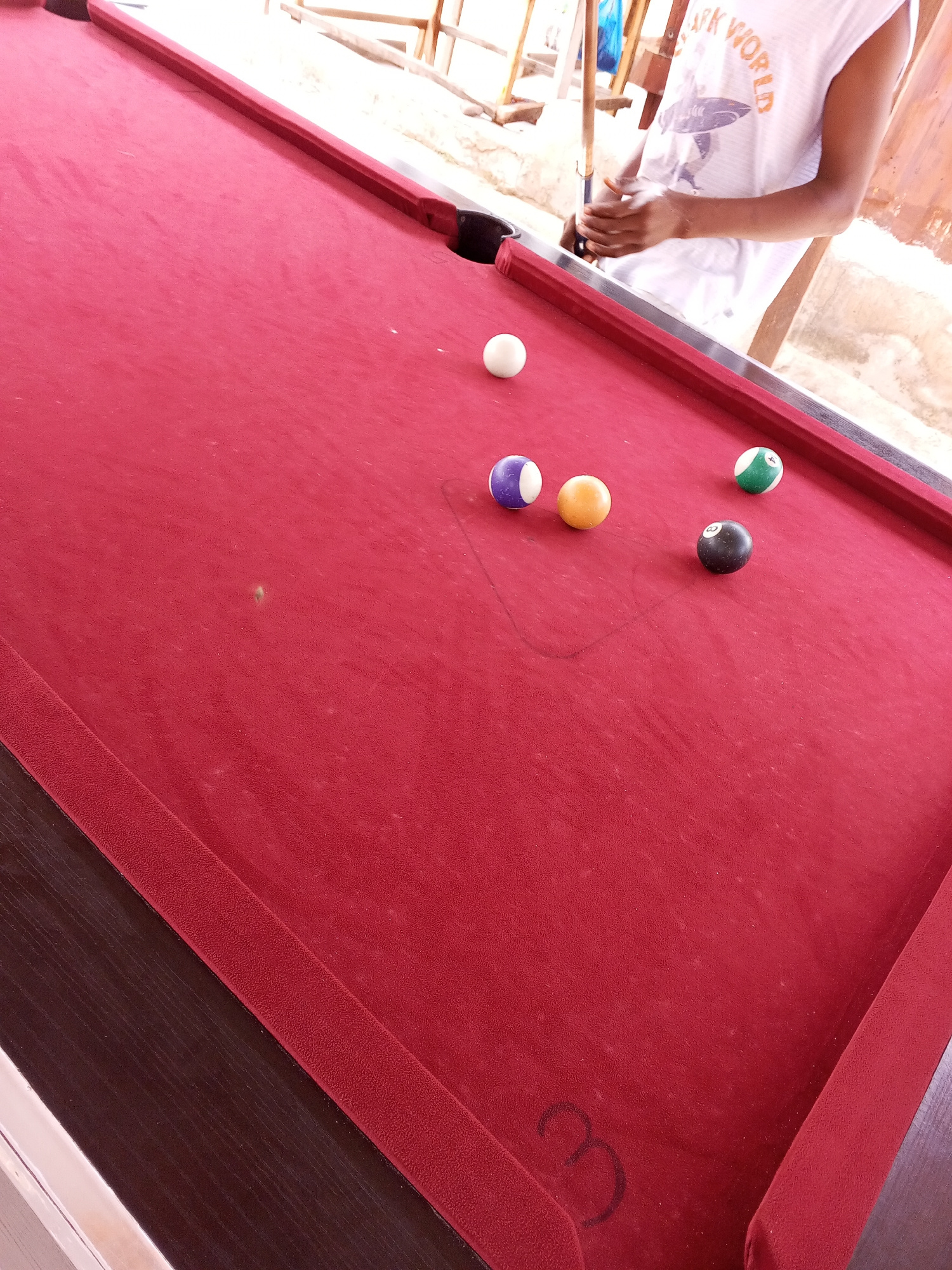 A high-angle shot shows a red pool table with several billiard balls scattered across its surface. A person, partially visible from the waist down, stands to the right, holding a pool cue in their hands, suggesting they are about to play or are in the process of a game. The background is blurred, but appears to be an outdoor setting with wooden structures, possibly indicating a casual establishment in Lokoja, Nigeria. The lighting is bright, suggesting daylight. The pool table appears to be in good condition, with a few minor scuffs or dust marks on the felt. The balls visible are a white cue ball, a purple and white striped ball, a yellow solid ball, a green and white striped ball with the number 4, and a black solid ball with the number 8.