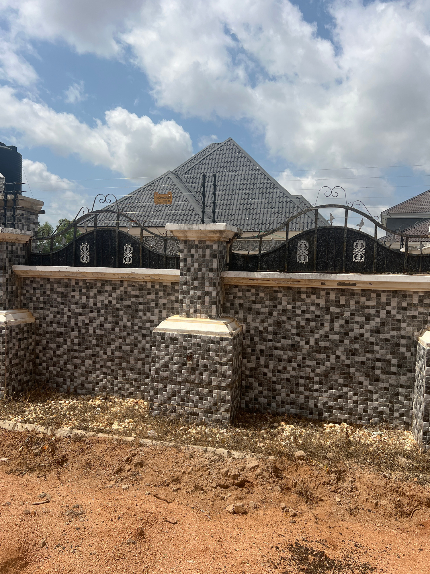 In Howayi, Nigeria, a tiled fence with ornate black iron gates stands before a house with a gray, tiled roof. The sky above is blue with scattered white clouds. In the foreground, the ground is covered in reddish-brown soil and dry, brown vegetation. A large black water tank is visible on the left side of the image, mounted on a support structure. The fence is constructed with alternating dark and light gray square tiles, and the gate posts are topped with white capstones. An orange sign with black lettering is affixed to the roof of the house. Electrical wires are strung across the scene, supported by poles. The scene appears to be a residential property on a sunny day.