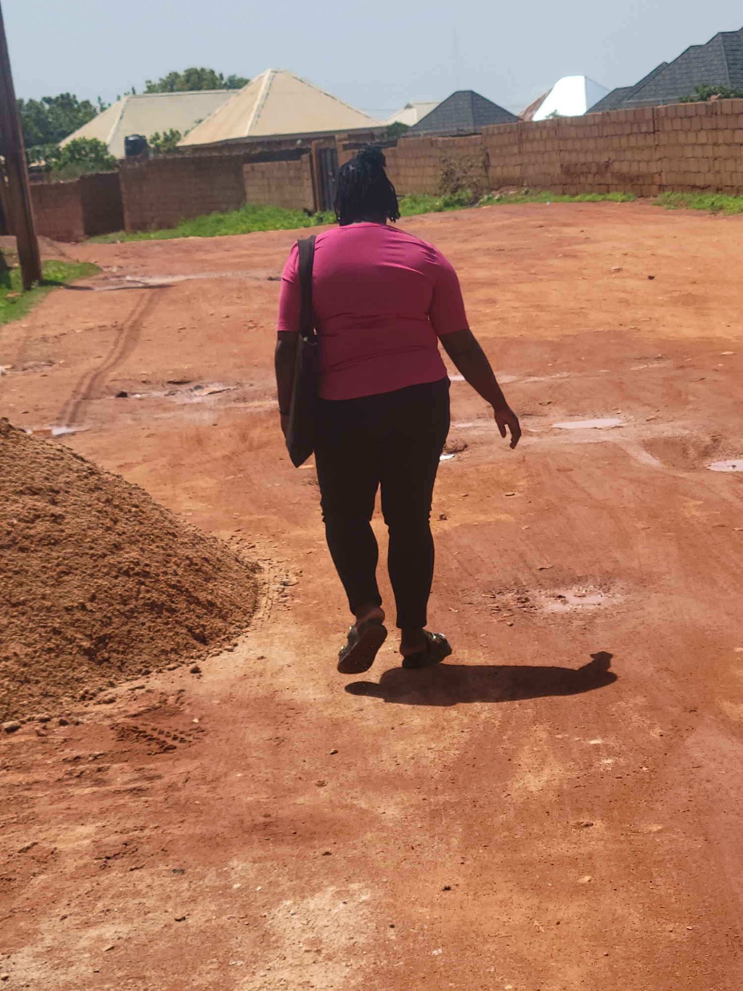 The image depicts a woman walking away from the camera on a dirt road in the city of Jos, Nigeria. She is wearing a bright pink t-shirt and black leggings, with a dark bag slung over her shoulder. Her hair is styled in braids. The road is uneven with puddles of water and tire tracks, indicating it may be a less developed area. To the left of the road, a large pile of sand is visible. In the background, several buildings with distinct architectural styles, including what appear to be newly constructed houses with corrugated iron roofs, are seen behind a low brick wall. The sky is clear and blue, suggesting a sunny day.