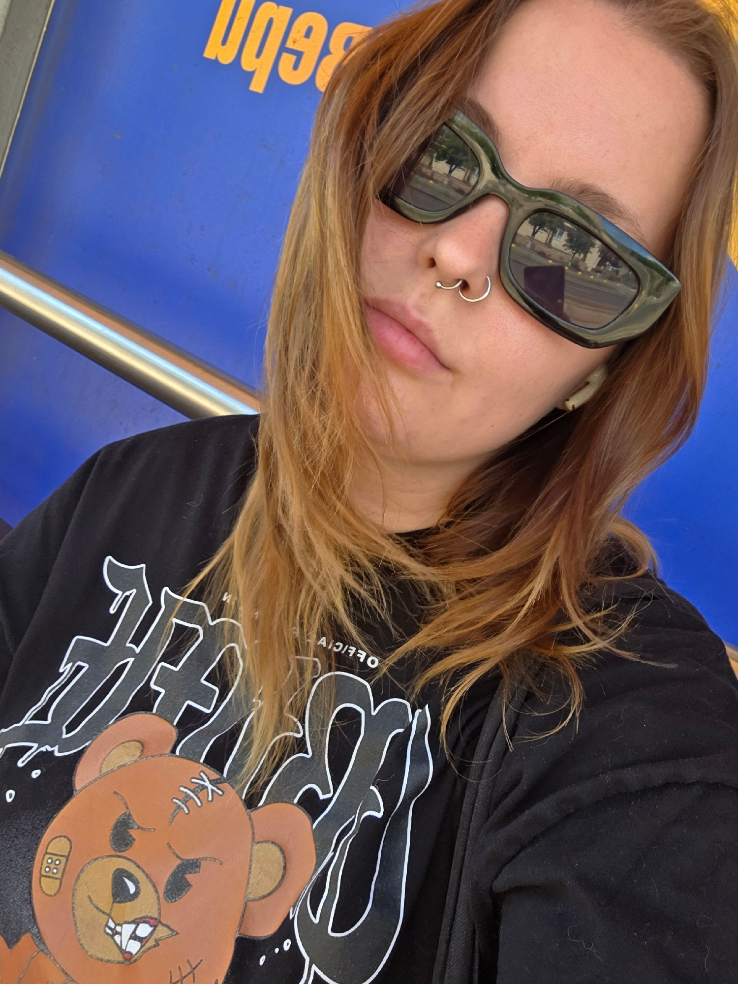 A young woman with auburn hair and a septum piercing is shown in a selfie taken outdoors. She is wearing dark-rimmed sunglasses and a black t-shirt with a graphic print of a teddy bear and stylized text. An earbud is visible in her right ear. She is seated in front of a bright blue surface with yellow text that appears to be part of an advertisement or signage, and a metal railing is present to her left. The background suggests an urban environment, potentially a public transit station or outdoor seating area. The lighting indicates it is daytime, with reflections of trees and buildings visible in her sunglasses. The setting is a street in the city of Minsk, Belarus, as suggested by the Cyrillic script on the blue background.