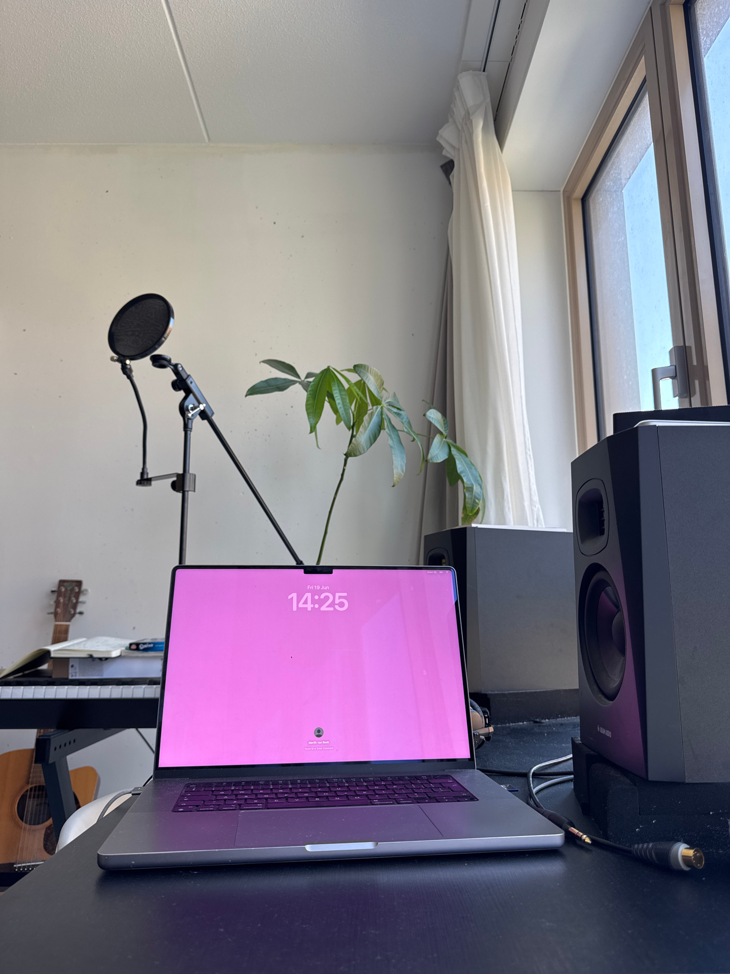 A home recording studio setup is depicted, oriented vertically. The scene features a desk with a laptop displaying a pink screen with the time "14:25" and "Fri 19 Jun". A microphone with a pop filter is mounted on a stand, positioned in front of a houseplant. In the background, two black speakers are visible, and a portion of a skylight is illuminated by daylight. The overall atmosphere suggests a creative workspace. A guitar is visible in the lower right corner. The provided location context is Amsterdam, Netherlands.