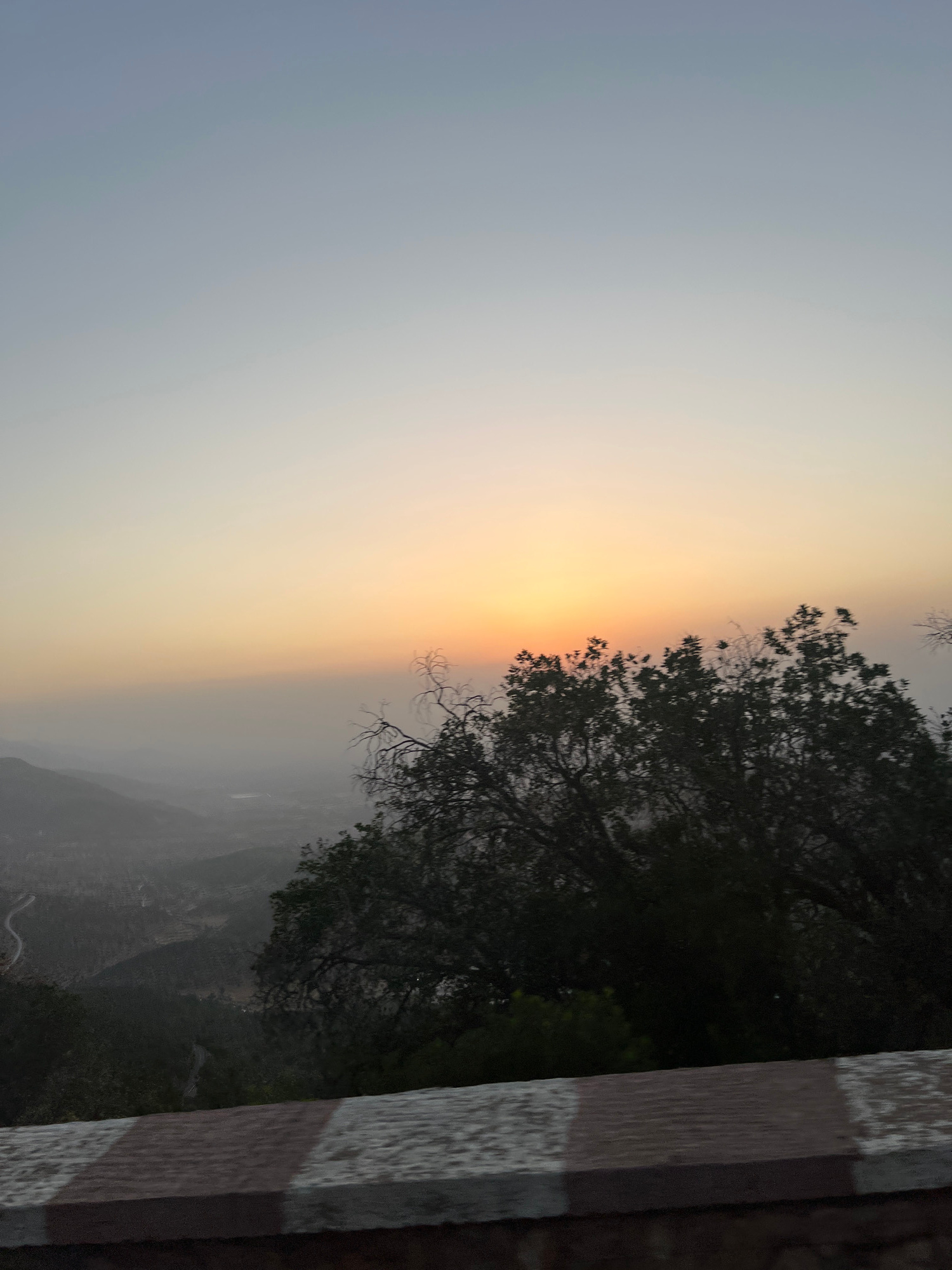 The image captures a serene sunset scene from a high vantage point overlooking a valley in Ouaouizert, Morocco. The sky transitions from a soft blue at the top to a warm orange and yellow glow near the horizon, indicating the setting sun. In the foreground, the dark silhouette of lush trees dominates the right side of the frame. The background reveals a hazy valley with rolling hills and what appears to be a winding road snaking through the landscape, suggesting a rural or mountainous area. A stone barrier with red and white stripes is visible at the very bottom, implying the viewpoint is from a roadside or overlook. There are no people or discernible actions depicted; the focus is on the natural landscape and the ambient light of the sunset. The overall impression is one of tranquility and natural beauty.