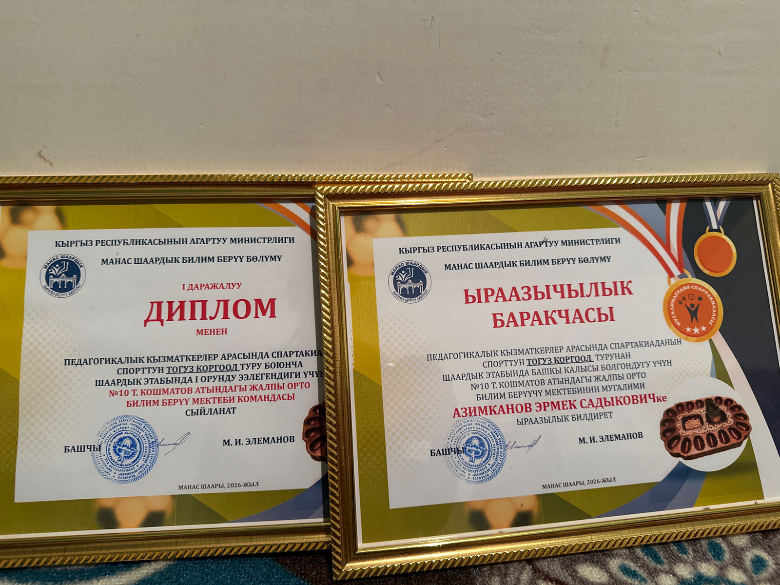 The image shows two framed certificates displayed on a patterned surface, possibly a carpet or upholstery. Both certificates are gold-bordered and feature text in what appears to be Kyrgyz. The certificate on the left is titled "ДИПЛОМ" (Diploma) and mentions " МЕНЕН" (with) below it. The certificate on the right is titled "БИРААЗЫЧЫЛЫК БАРАКЧАСЫ" (Certificate of Achievement) and also includes text related to sports and education. Both certificates have seals and signatures. A medal is visible in the upper right corner of the right certificate. The lighting suggests an indoor setting. The location is identified as Ленин айылы, Kyrgyzstan.
