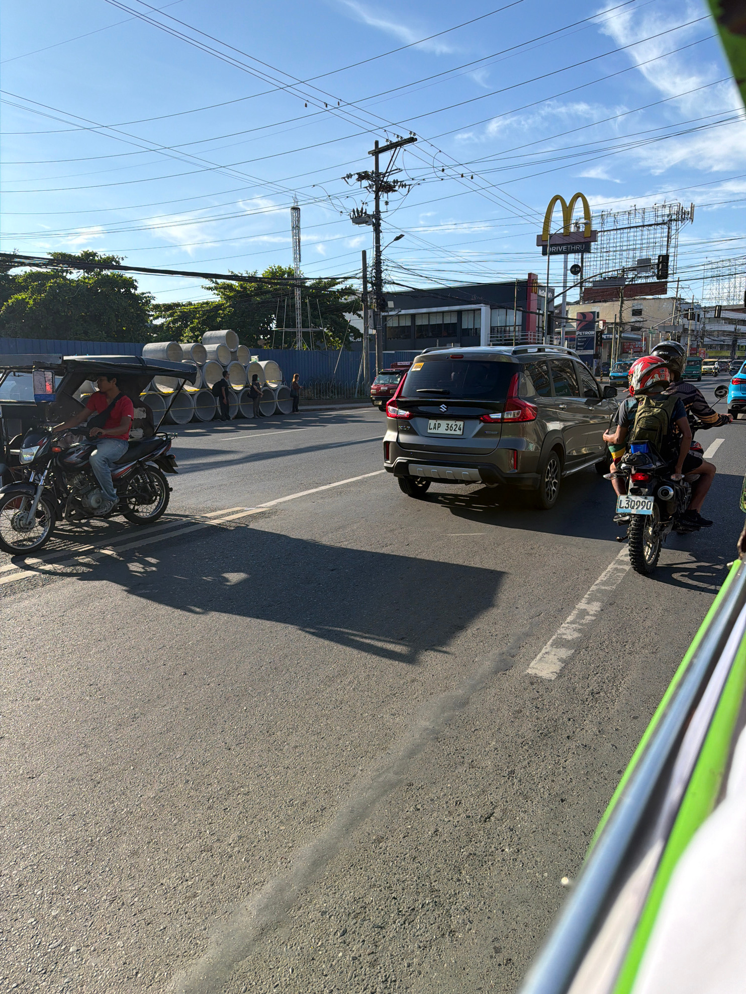 This image captures a street scene in Davao City, Philippines, under a clear blue sky with scattered clouds. The road is paved and appears to be a multi-lane thoroughfare. In the foreground, a motorcycle with a sidecar is stopped, and the driver, a man wearing a red shirt and blue jeans, is seated. Further ahead, a grey SUV with the license plate "LAP 3624" is in motion. Behind the SUV, another motorcycle carrying two individuals, one wearing a red helmet, is also moving.
On the right side of the road, a building with a "McDonald's Drive-Thru" sign is visible. Several utility poles with a complex network of wires are present throughout the scene. To the left, a construction area is indicated by a blue fence and a stack of large concrete pipes. Green trees line the background, interspersed with buildings and more overhead wires. The overall impression is a typical urban environment in the Philippines, with ongoing traffic and commercial establishments.