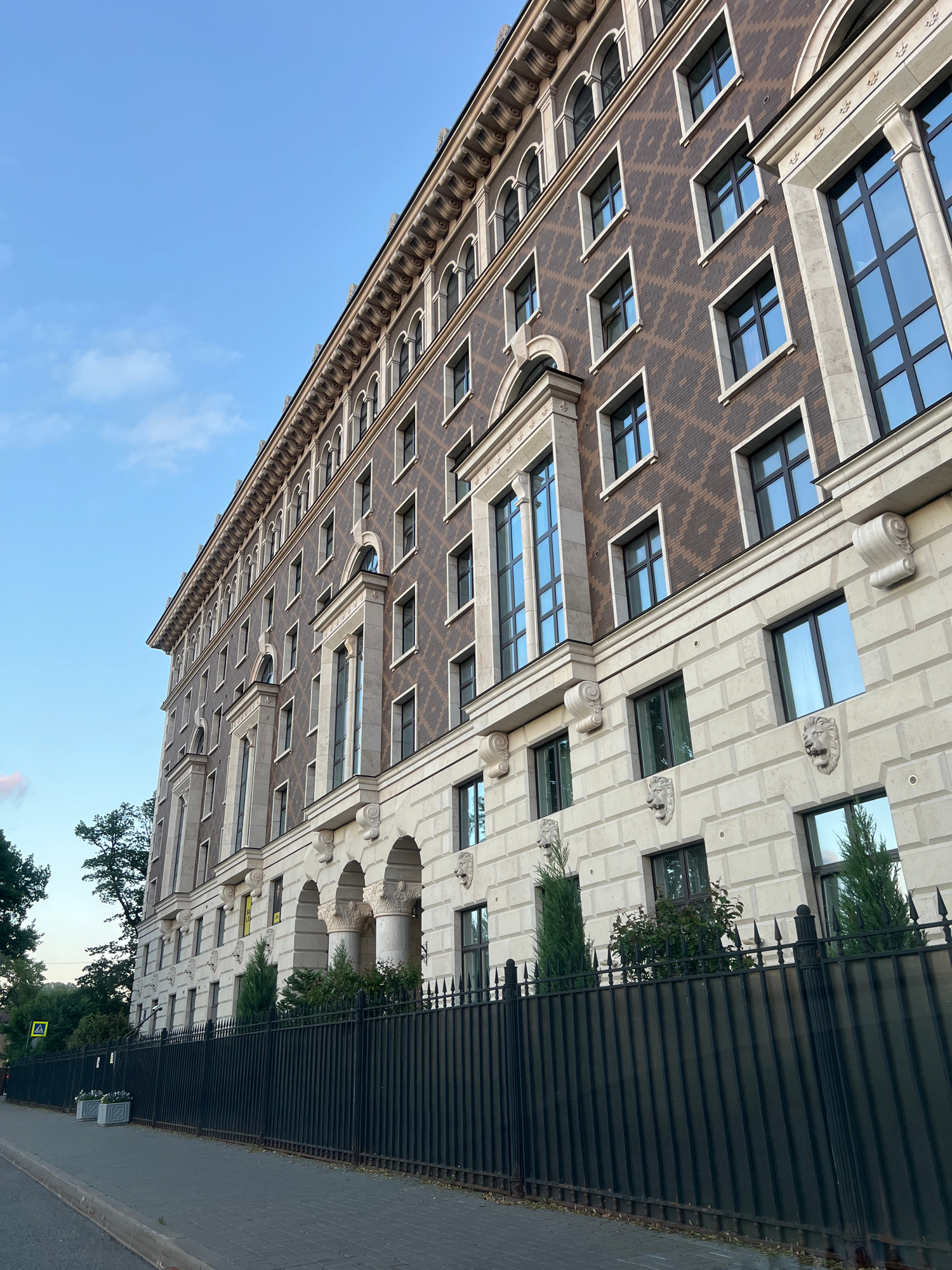 The image shows a long, multi-story building with a classical architectural style. The building has a dark brown facade with a repeating diamond pattern and is adorned with light-colored stone accents, columns, and decorative lion heads. There are numerous windows, some arched and some rectangular, with dark frames. A black metal fence runs along the front of the building, separating it from a paved sidewalk and a street. Trees and greenery are visible behind the fence and on the left side of the image. The sky is clear with a few wispy clouds. This building is located at 10 Krestovsky Prospect in Saint Petersburg, Russia. There are no people or vehicles visible in the frame.