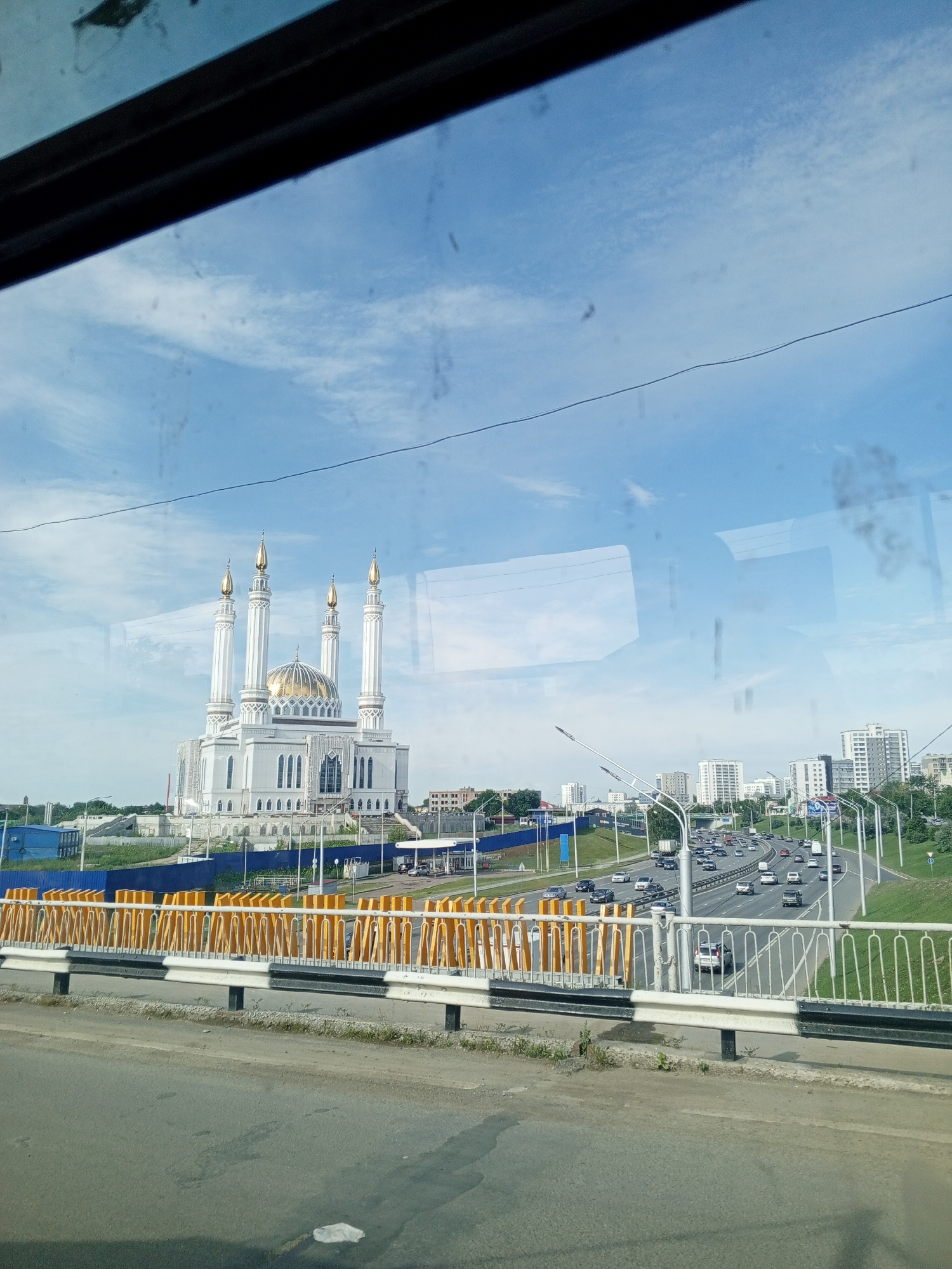 The image shows a view from a bridge overlooking a highway in Ufa, Russia, with a prominent mosque in the background. The mosque, identified as the L'ala-Tul'pan Mosque, is a large, white building with a golden dome and four tall minarets topped with golden spires. The mosque is situated on a hill, with a city skyline of modern buildings in the distance. The highway below is busy with traffic, with cars and trucks visible on multiple lanes. The bridge itself has a white railing with black and yellow stripes on its lower section. The sky is blue with scattered white clouds. The overall scene depicts a contemporary urban landscape with a significant religious landmark.