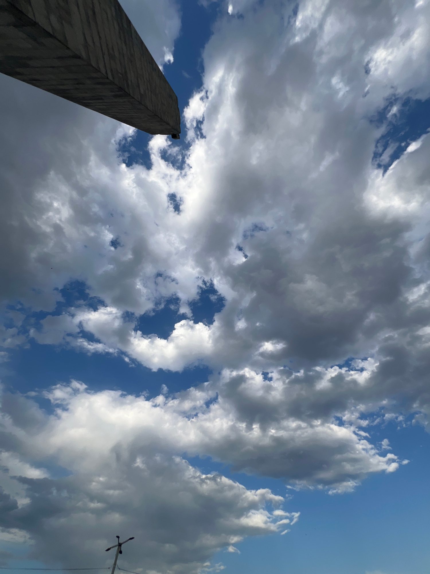 The image shows a view from below, looking up at a dramatic sky filled with a mix of light and dark clouds against a bright blue sky. In the lower left of the frame, a tall, dark, stone obelisk dominates the scene, angled upwards and piercing the clouds. The obelisk has a rough, textured surface, suggesting it is made of stone blocks. The sky is dynamic, with fluffy white clouds interspersed with darker, more ominous storm clouds, creating a sense of movement and depth. The sunlight filtering through the clouds suggests it is daytime, possibly late afternoon given the contrast between light and shadow. On the far right of the image, a faint silhouette of what appears to be a lamppost or antenna can be seen against the sky. The overall mood is one of grandeur and perhaps a touch of foreboding due to the cloudy sky. Without further context, the specific location cues are limited to the obelisk and the sky. Based on the provided location context, this obelisk could be a monument in Yerevan, Armenia. There is no visible text in the image.