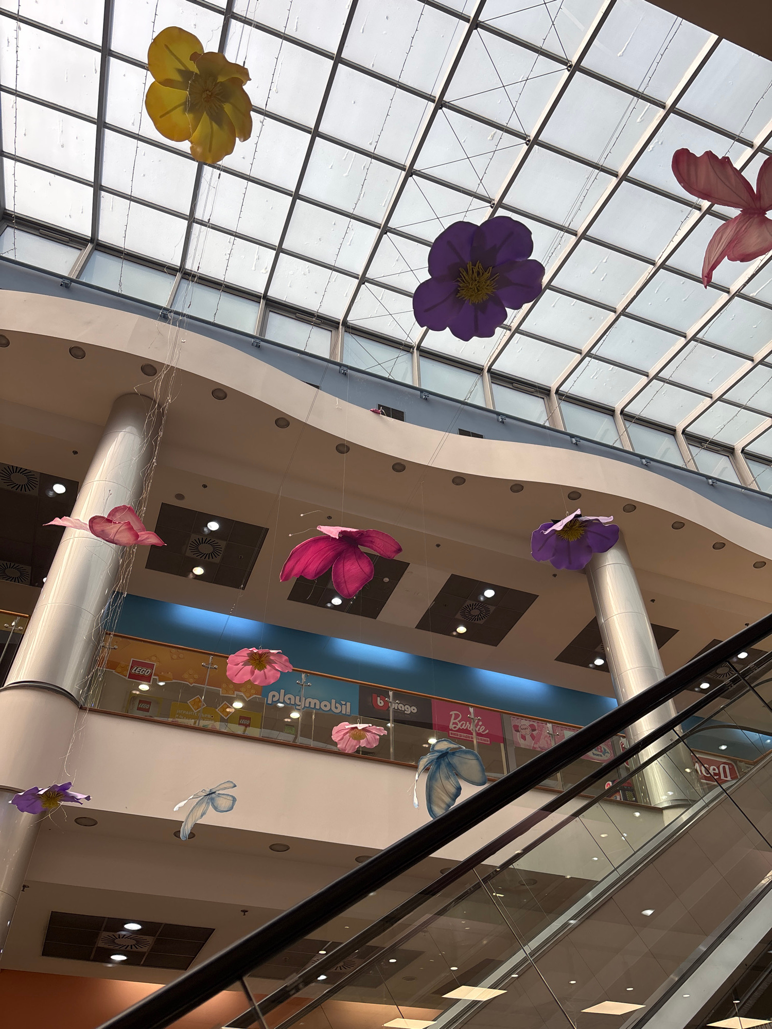 This media file captures an interior view of a shopping mall, looking upwards towards a large, angled glass ceiling. The scene is decorated with large, colorful artificial flowers and butterflies suspended from the ceiling by thin wires, creating a whimsical and festive atmosphere. The flowers are in various shades of purple, pink, and yellow, and some appear to be made of fabric. A blue butterfly decoration is also visible, adding to the playful theme.
The mall's architecture features curved, cream-colored walls and large cylindrical pillars. The glass ceiling is divided into a grid of panes, allowing natural light to filter in. Below the ceiling, various levels of the mall are visible, including escalators and shop fronts.
Several shop signs are partially visible, including "Playmobil" and "Lego." One sign features the text "Barbie" prominently. The lighting within the mall suggests it is daytime, though the exact time of day cannot be determined. The overall mood is bright and inviting, indicative of a commercial space designed for leisure and shopping. The location is identified as Pleven, Bulgaria. There are no people visible in the frame.