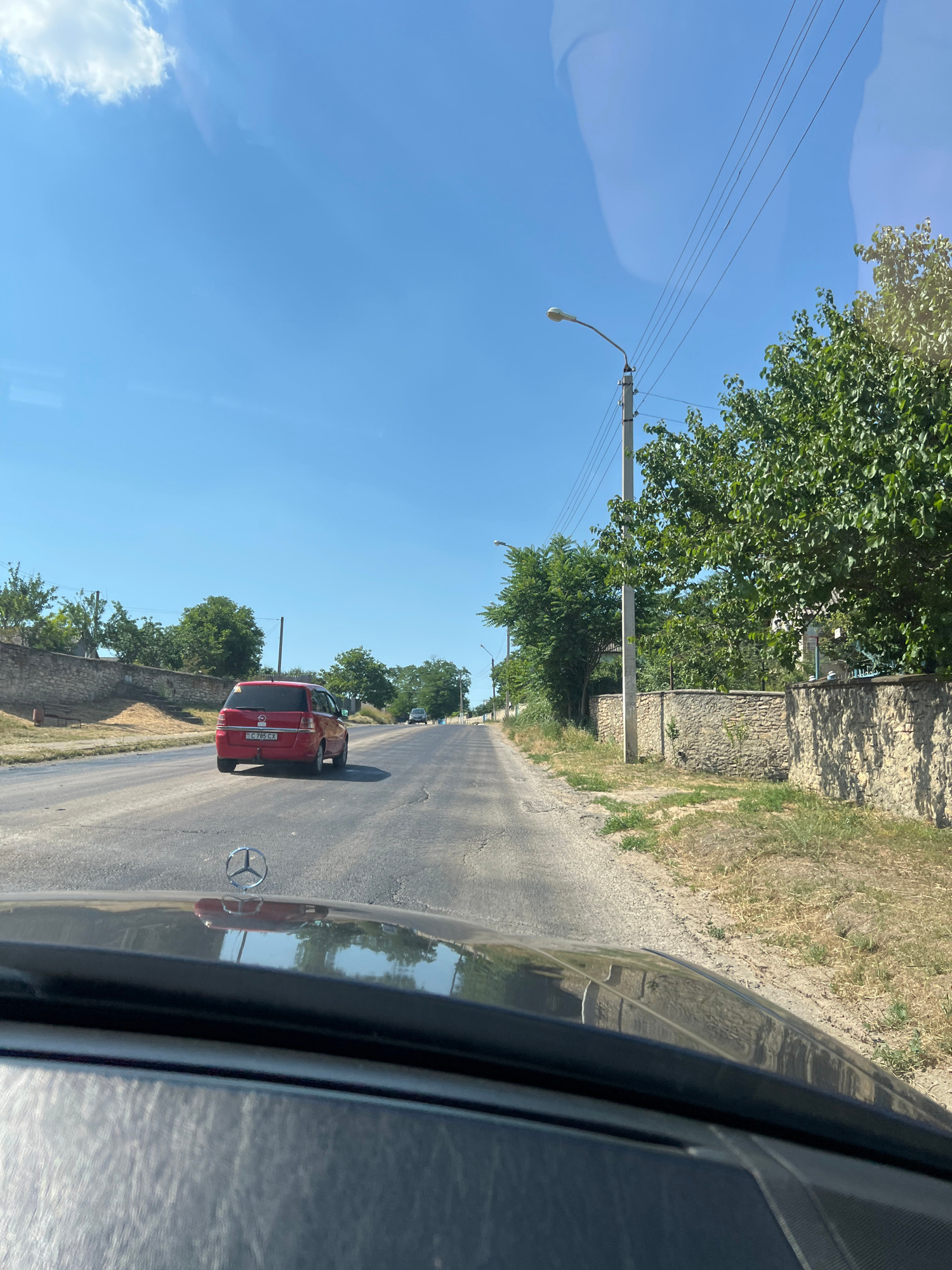 A red minivan is driving down a paved road on a sunny day. The minivan has a license plate that reads "G C802 CX". The road is bordered by a dry, grassy area and a stone wall on the left, and trees and a stone wall on the right. Overhead, power lines and a street lamp are visible. The sky is clear blue with a few wispy clouds. The photo appears to have been taken from inside another vehicle, as the side of a car and its reflection are visible in the foreground. The scene suggests a rural or semi-rural setting, possibly in Tashlyk, Moldova, given the location context. The time of day appears to be late morning or early afternoon due to the bright sunlight.