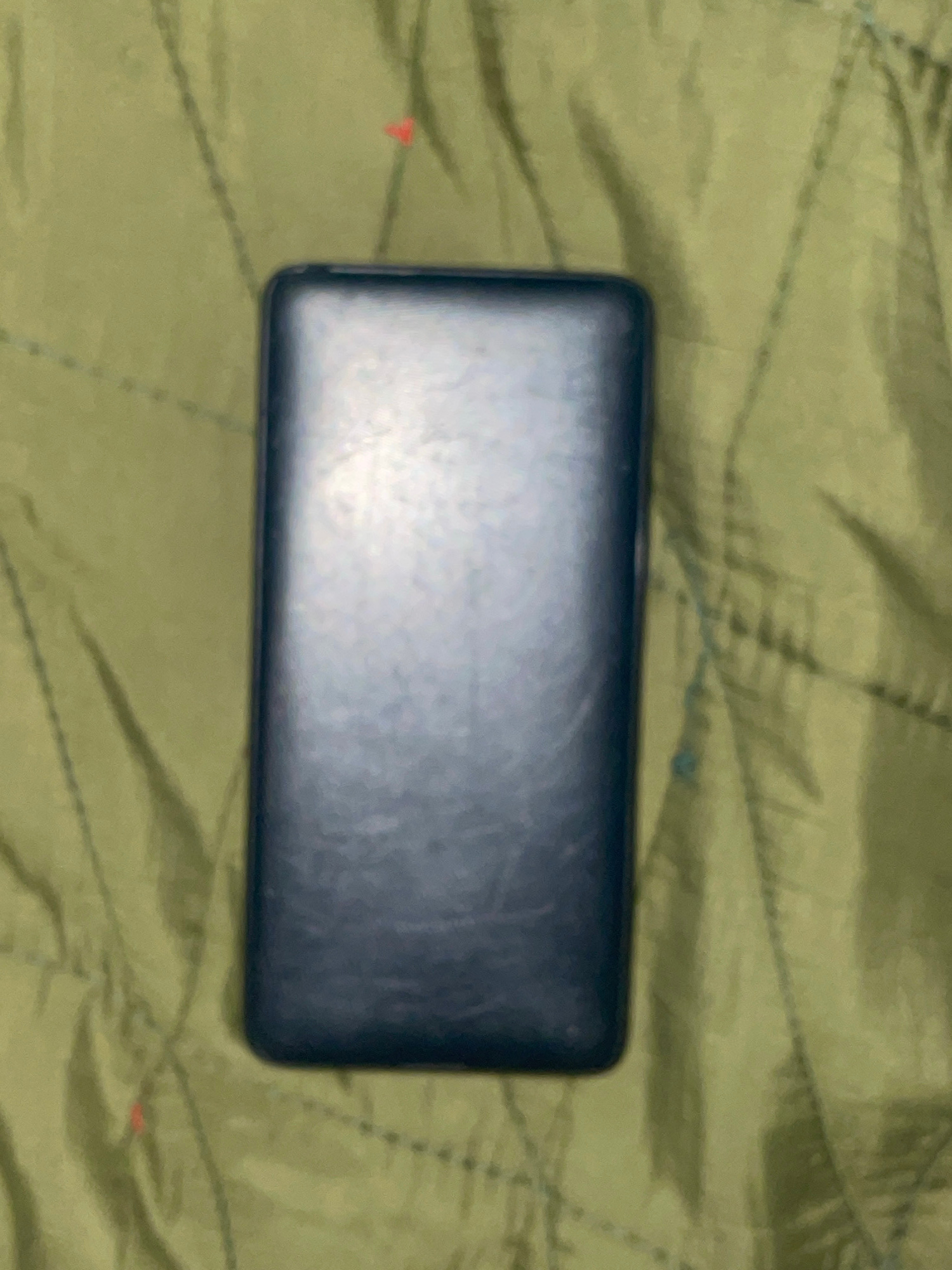 This is a medium-shot, overhead photograph of a dark, rectangular object resting on a textured, olive-green surface. The object appears to be a phone or a similar electronic device, lying horizontally in the center of the frame. Its surface is smooth and shows some subtle reflections, suggesting it might be made of glass or plastic. The green background has a quilted or padded texture with a pattern of diagonal lines and occasional darker shadows. There are a few small, indistinct red and blue marks scattered on the green surface, possibly minor blemishes or color variations. The lighting in the image is somewhat diffused, creating soft shadows and a slightly muted color palette. The overall scene is static, with no apparent activity or human presence. The context of Jalingo, Nigeria, is provided, but there are no specific visual cues within the image to confirm this location. The time of day and weather are not discernible. No text is visible.