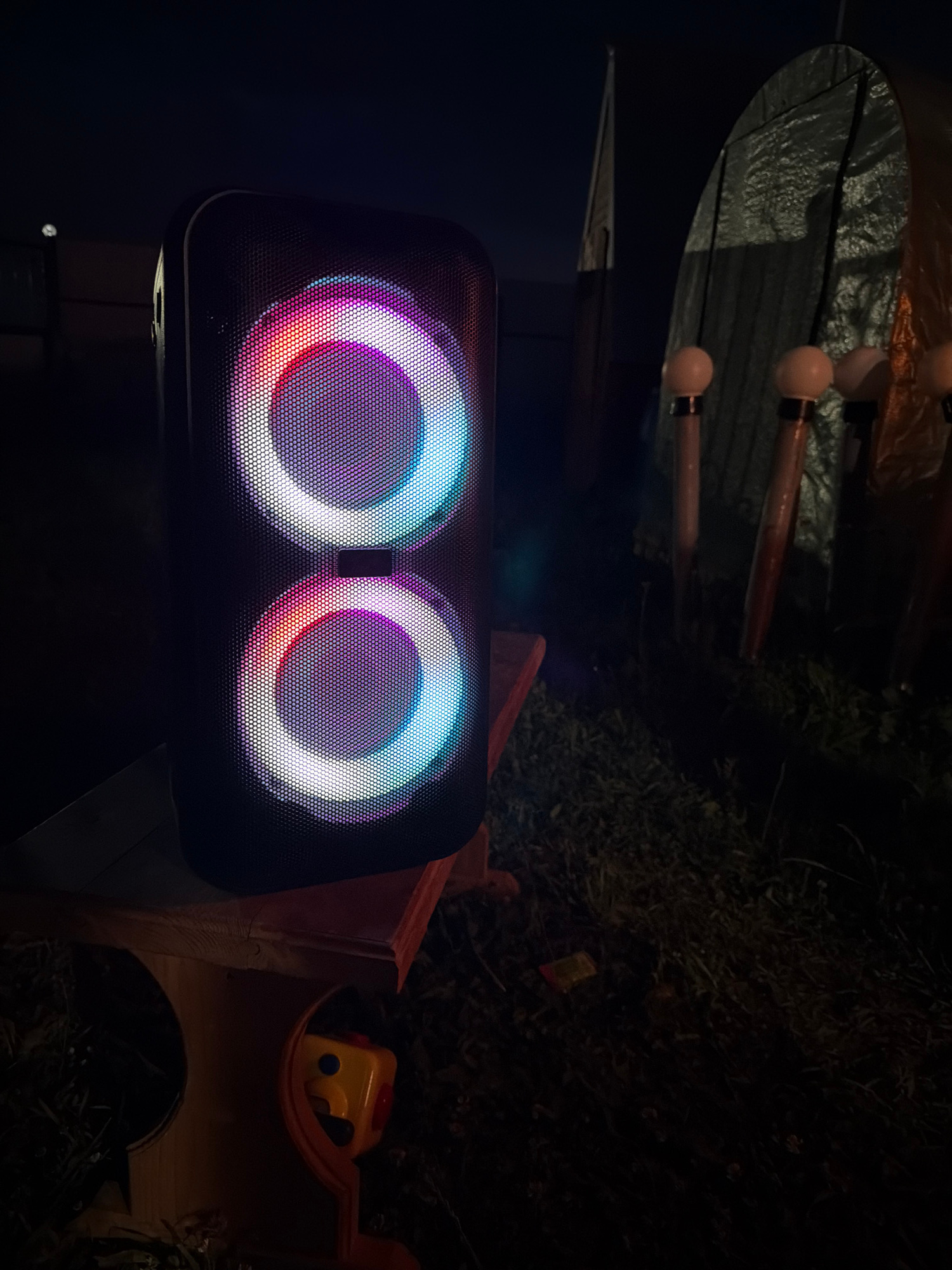 A black, rectangular speaker with two illuminated circular grills sits on a wooden bench in a dark outdoor environment. The grills emit a colorful glow, transitioning from red and pink on the left to blue and white on the right. Behind the speaker, several long, thin objects with spherical tops stand upright in the shadows. The ground is covered in grass and debris. A portion of a tarp or tent is visible in the background. A small, yellow toy with a red button is partially visible on the bench. The scene appears to be a nighttime setting.
