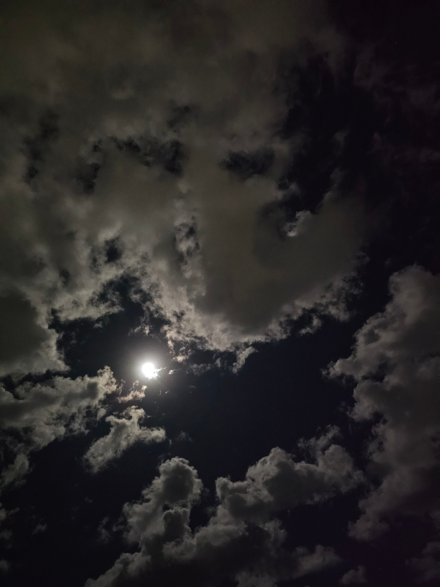 This is a photograph of the night sky, taken from Denpasar, Indonesia. The image is dominated by dark, textured clouds illuminated by the bright light of the moon, which is visible as a luminous disc peeking through a gap in the clouds. The clouds are varied in density and shape, with some appearing wispy and translucent, while others are thick and opaque, creating dramatic contrasts of light and shadow. The overall impression is one of a dynamic, cloudy night.