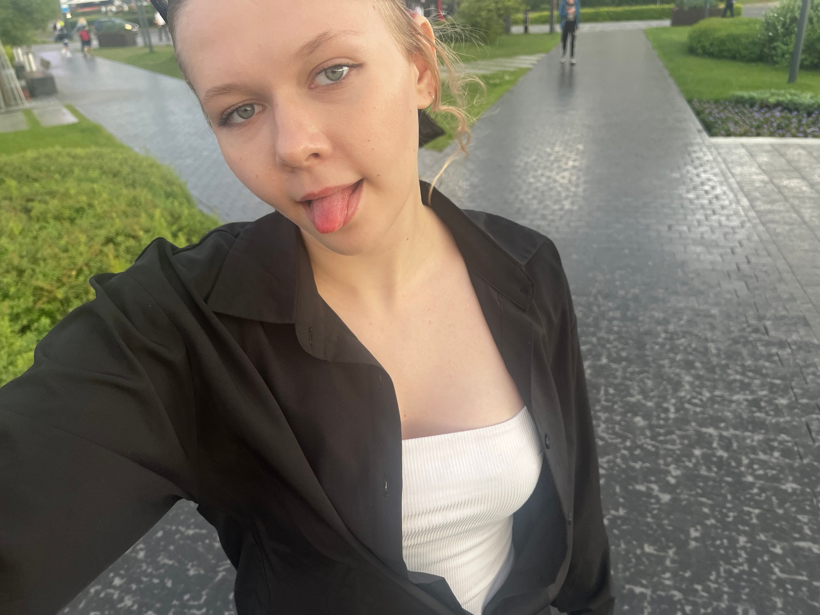 A young Caucasian woman with blonde hair tied back and her tongue sticking out is taking a selfie outdoors. She is wearing a black open button-up shirt over a white ribbed tank top. The background shows a paved walkway with cobblestone patterns, green grass and bushes, and some people walking in the distance. The overall setting appears to be a park or public walkway in the city of Perm, Russia.