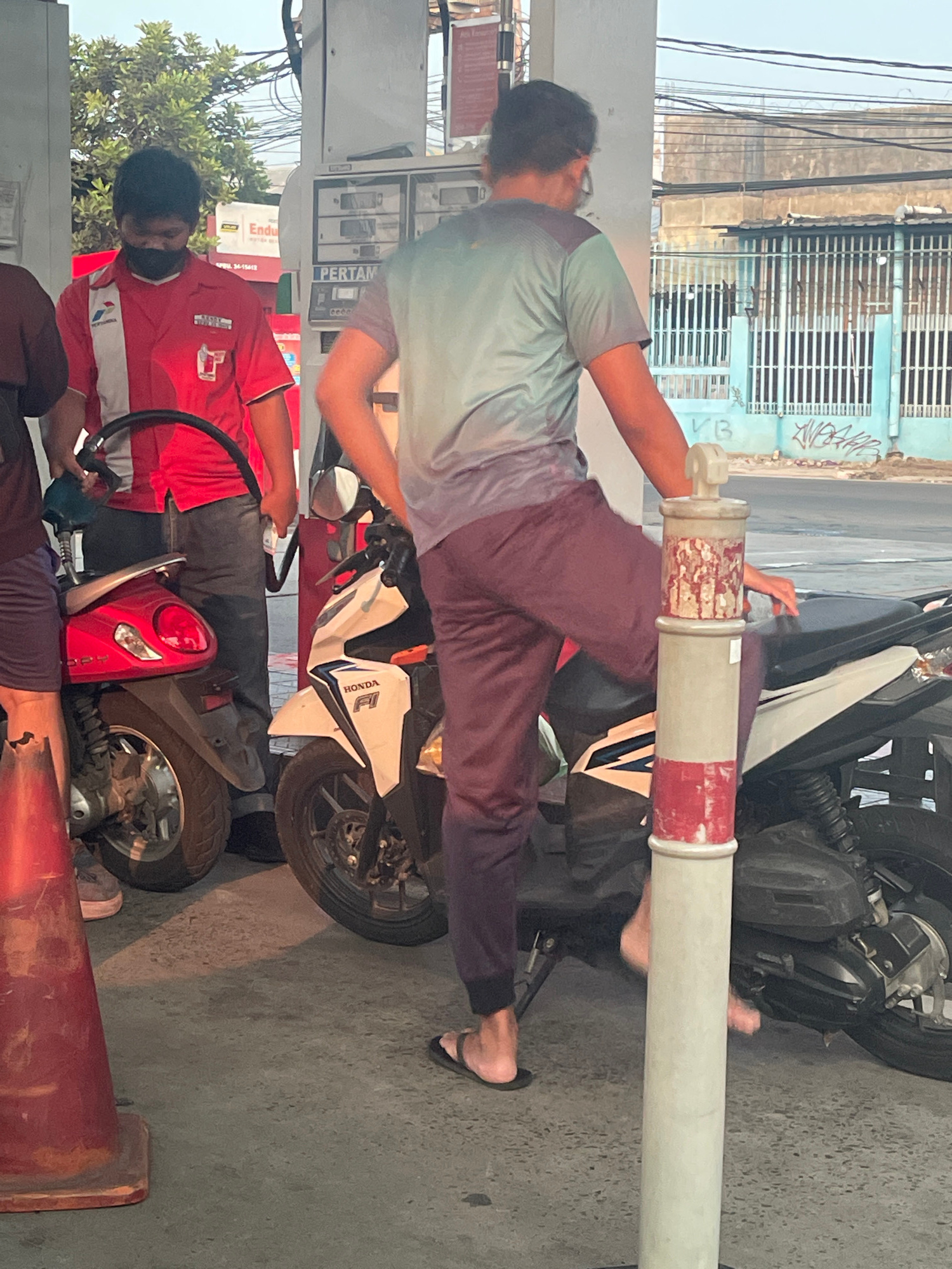 A gas station attendant in a red uniform with a black face mask is standing next to a motorcycle, holding a gas nozzle. Another person in a teal shirt and maroon pants is also near a motorcycle. The scene appears to be a Pertamina gas station in South Tangerang, Indonesia, with other motorcycles and a traffic cone visible. The background shows buildings and a street.