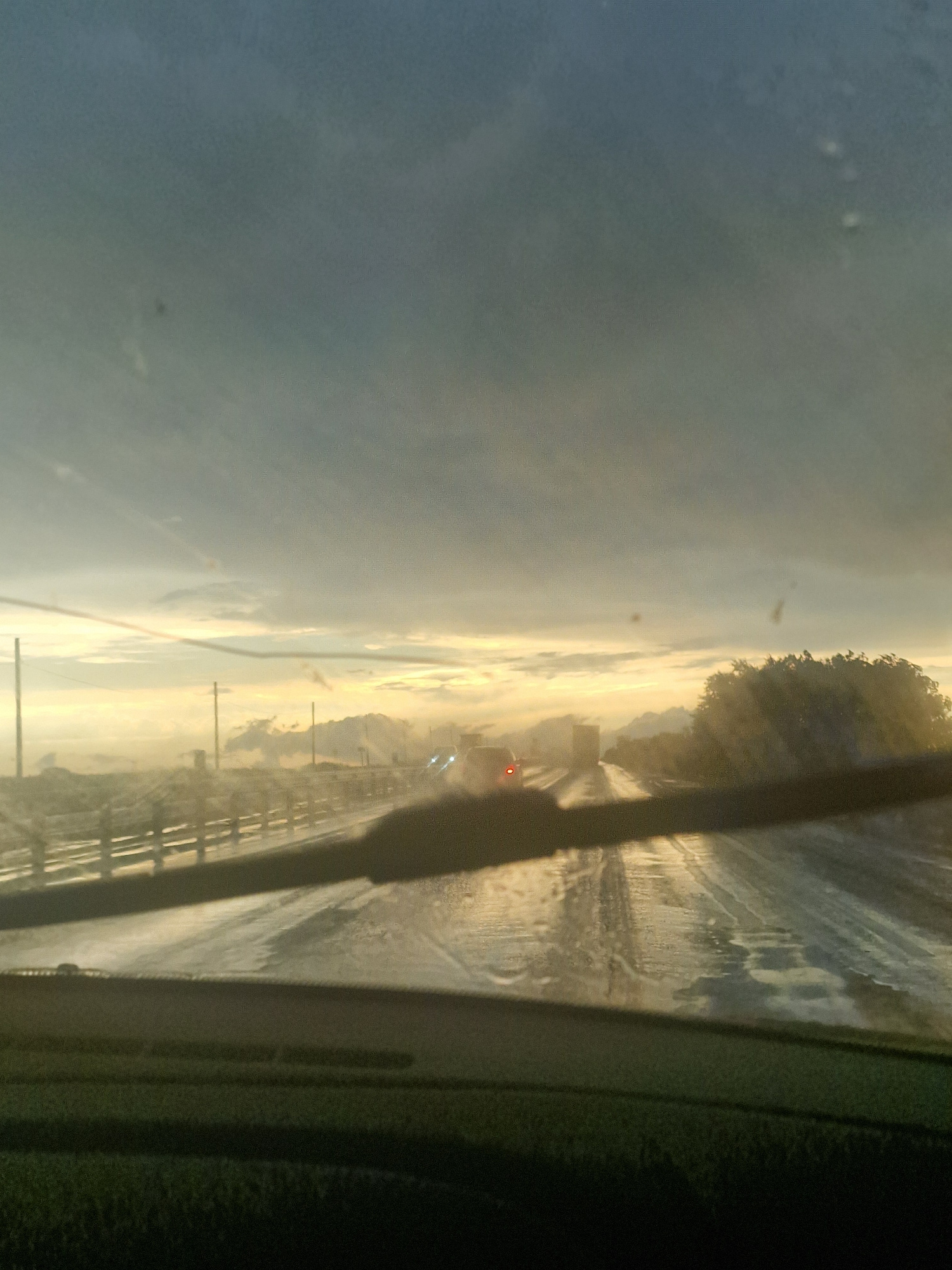The image captures a view from inside a vehicle, looking outwards through the windshield. The perspective is tilted, giving the impression of motion or an unusual camera angle. The scene appears to be a roadside, likely in Russia, given the location context.
The primary subjects are the road and the sky. The road is wet, reflecting the light and suggesting recent rain or moisture. There are tire tracks visible on the road surface. Along the side of the road, there is a fence-like structure, possibly a guardrail or a field boundary. In the distance, there is a tree line, with some foliage visible. A single red light, possibly from another vehicle, is noticeable on the road ahead.
The sky is overcast with a hazy quality, indicating diffused light. The overall lighting suggests it might be dawn or dusk, as the sky has a soft, luminous glow. The weather appears to be dull or possibly misty, contributing to the muted colors and the wetness of the road.
There is no visible text in the image. The emotion conveyed is one of movement and possibly introspection, given the solitary perspective and the atmospheric conditions. The scene is characteristic of driving through a rural or semi-rural area, potentially on a highway or a main road.