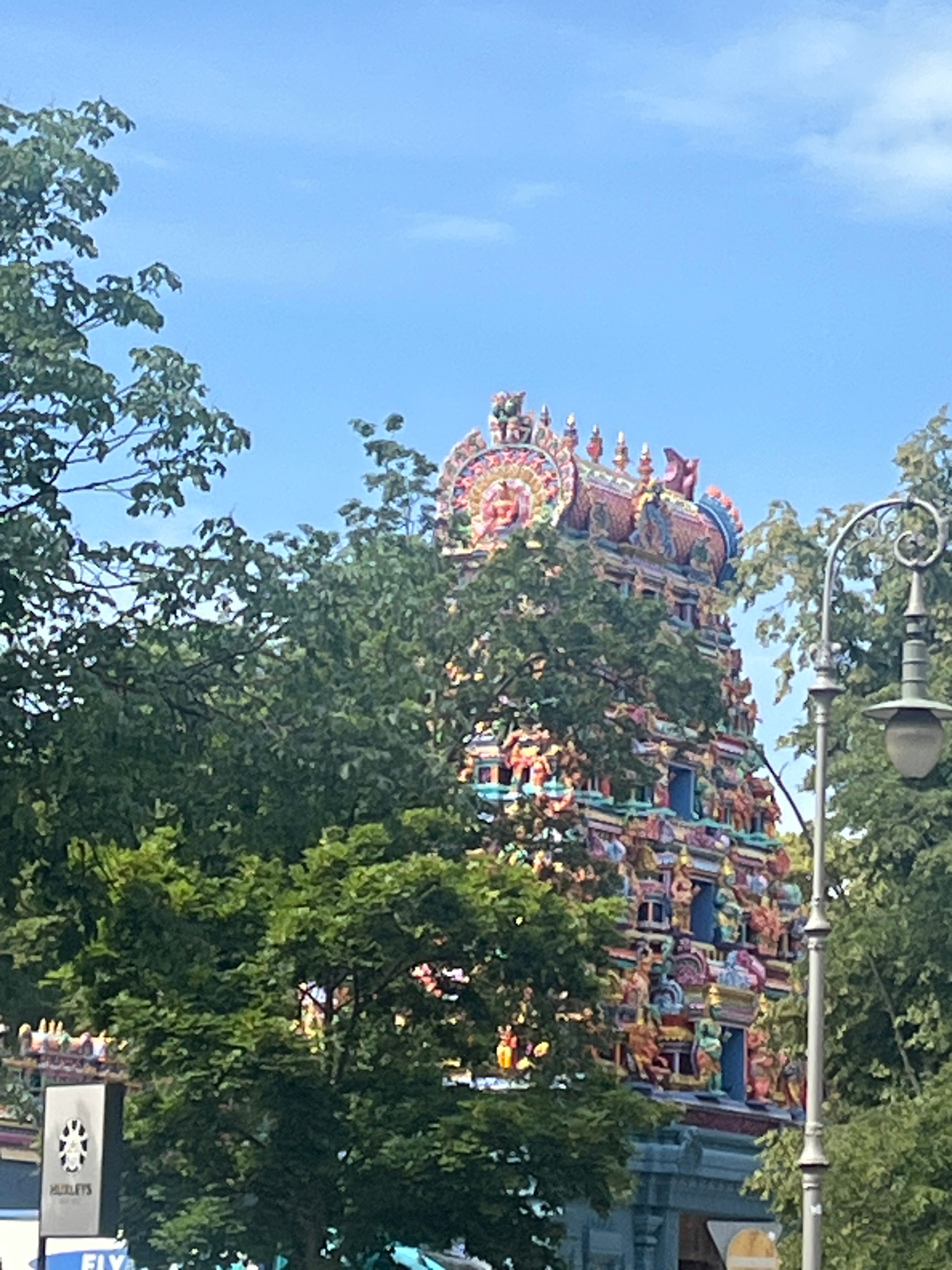 This image captures the vibrant and intricately decorated tower of a Hindu temple, partially obscured by lush green trees, set against a clear blue sky. The temple's facade is adorned with colorful sculptures of deities and mythological figures, reflecting a rich artistic and cultural heritage. The overall scene suggests a peaceful, bright day in a city environment. Based on the distinctive architecture of the Sri Radha Krishna Temple, this image was taken in Berlin, Germany.