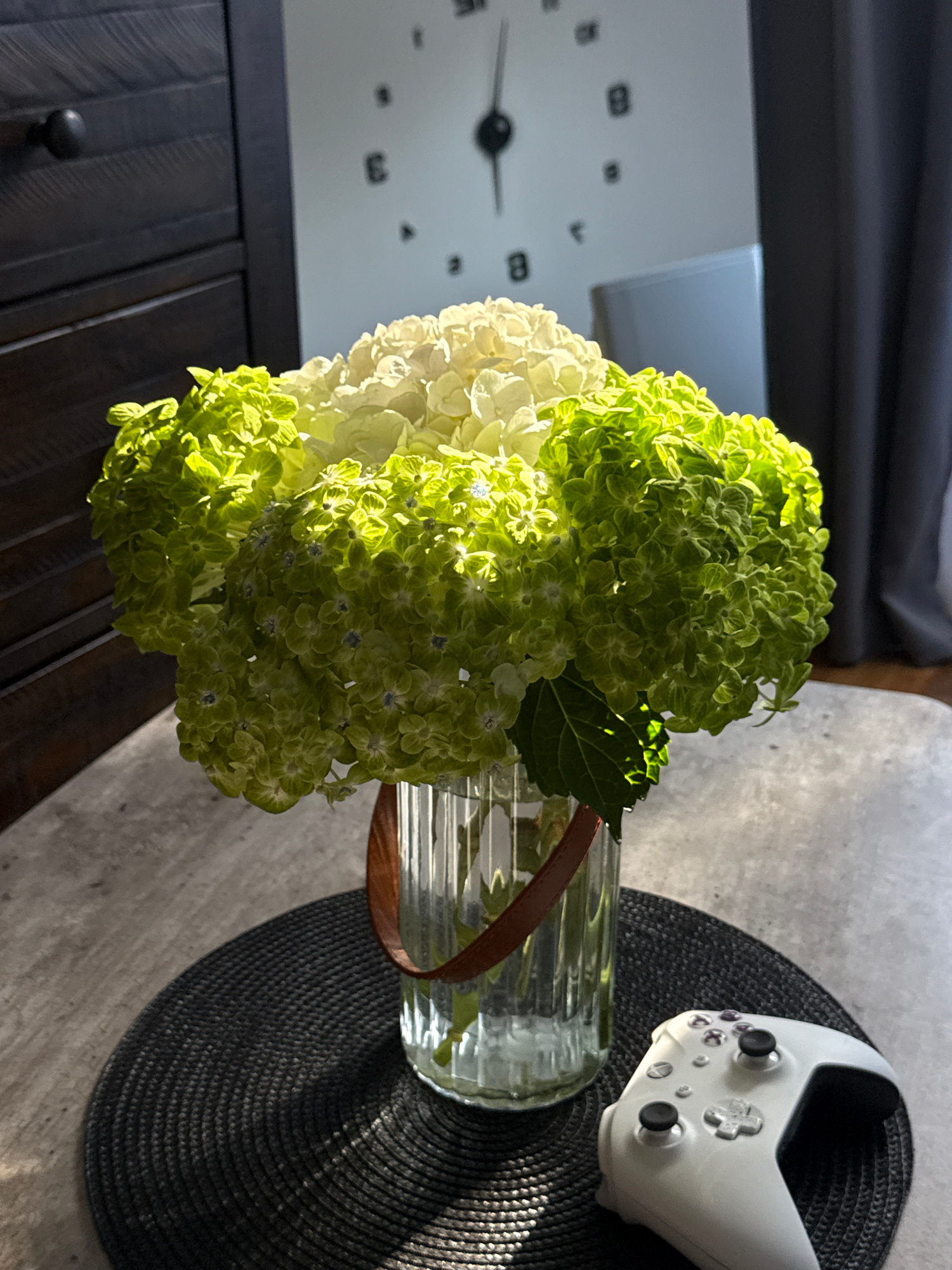 A white Xbox controller rests on a dark circular placemat on a stone-patterned table. Behind it, a clear glass vase holds a large bouquet of white and green hydrangeas. The vase has a brown leather strap around its neck. In the background, a wall clock with a minimalist design is visible, along with dark wooden furniture and grey curtains. The scene is lit by natural light, casting shadows on the table and flowers.