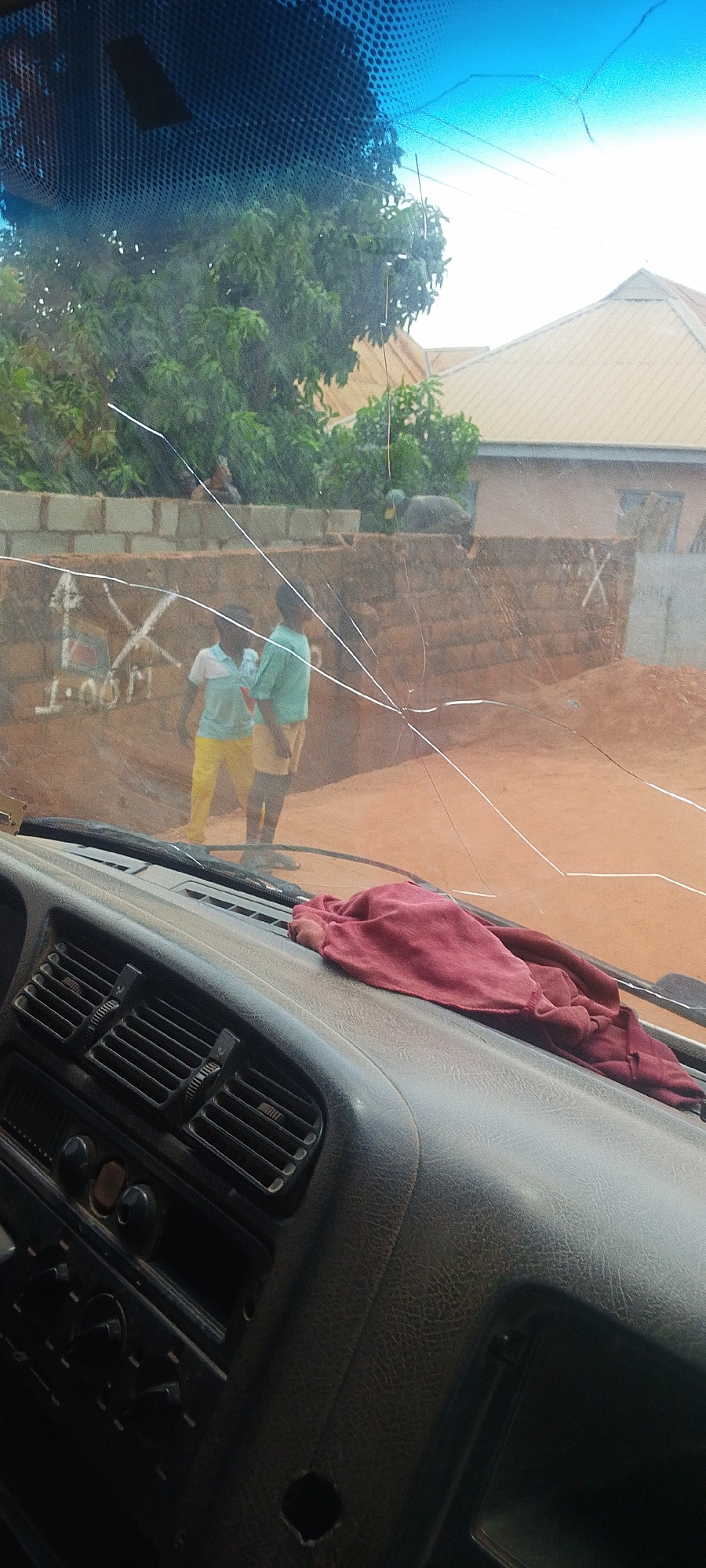 The image captures a scene viewed through a cracked car windshield, likely from inside a vehicle in Jos, Nigeria. The windshield has multiple prominent cracks obscuring the view. In the background, two young boys are visible standing near a brick wall. One boy wears a light blue and white striped shirt and yellow pants, while the other wears a light blue t-shirt and light brown shorts. The wall behind them is constructed of reddish-brown bricks and has some markings, including what appears to be an 'X' and some unreadable text. Beyond the wall, trees with green foliage and a building with a light-colored roof are visible, suggesting an outdoor environment within a populated area. In the foreground, the interior of the car is visible, including the dashboard with air vents and a dark red cloth draped over it. The ground in front of the wall appears to be reddish-brown earth. The overall impression is a casual outdoor setting, possibly a street or an open area adjacent to a residential or construction site in Jos.
