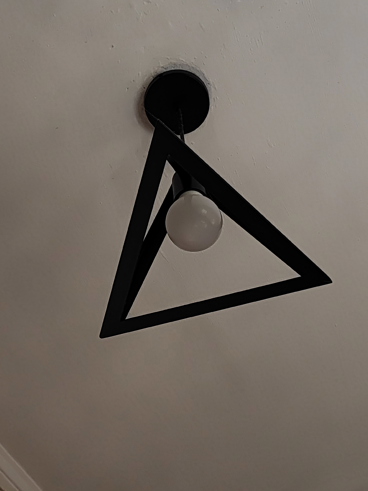 The image shows a modern black triangular ceiling light fixture with a white bulb, mounted on a textured white ceiling. The fixture is the primary subject, positioned in the center of the frame. The ceiling appears to have some minor imperfections or textures. No people or other significant objects are visible. The lighting is soft, suggesting an indoor environment. The photograph was taken in the city of Korosten, Ukraine.